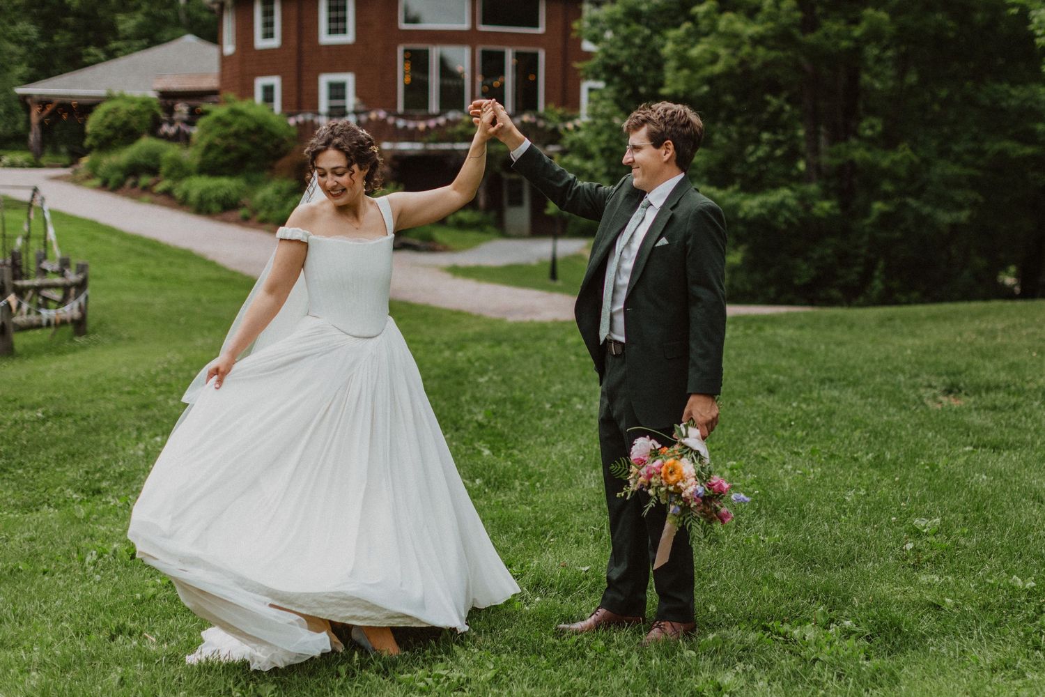 Bride and groom dancing on green lawn outside brick estate, groom spinning bride in white gown while holding colorful bouquet.