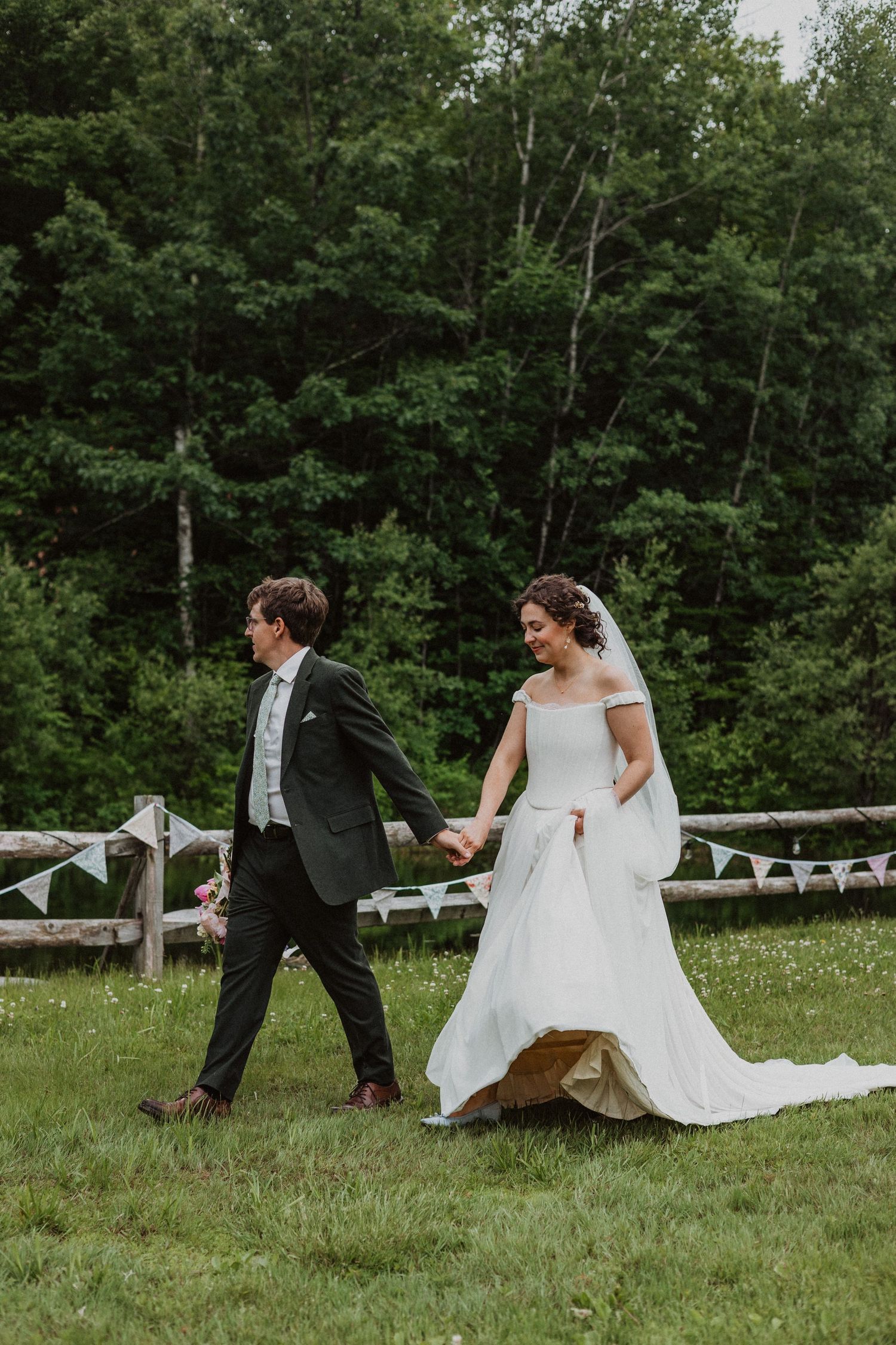 Bride and groom walking hand in hand across green meadow with rustic fence and forest backdrop.