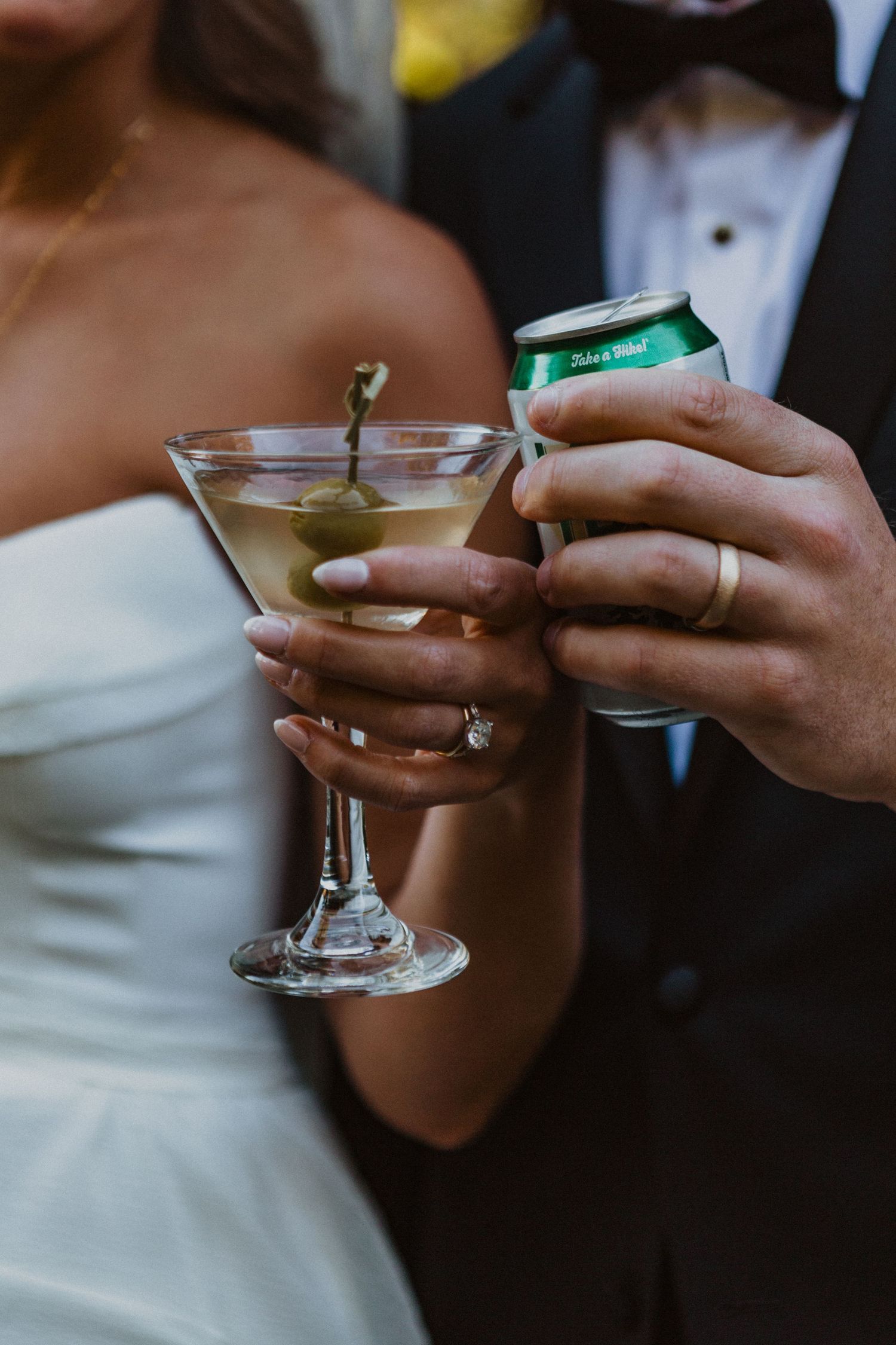 Bride holding martini with olive and groom holding beer can, both wearing wedding rings, toasting at their reception.
