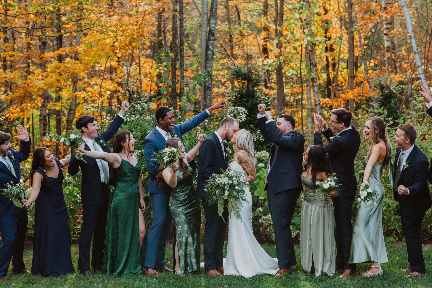 Wedding party celebrating with kiss in vibrant fall foliage forest wearing green and navy attire.