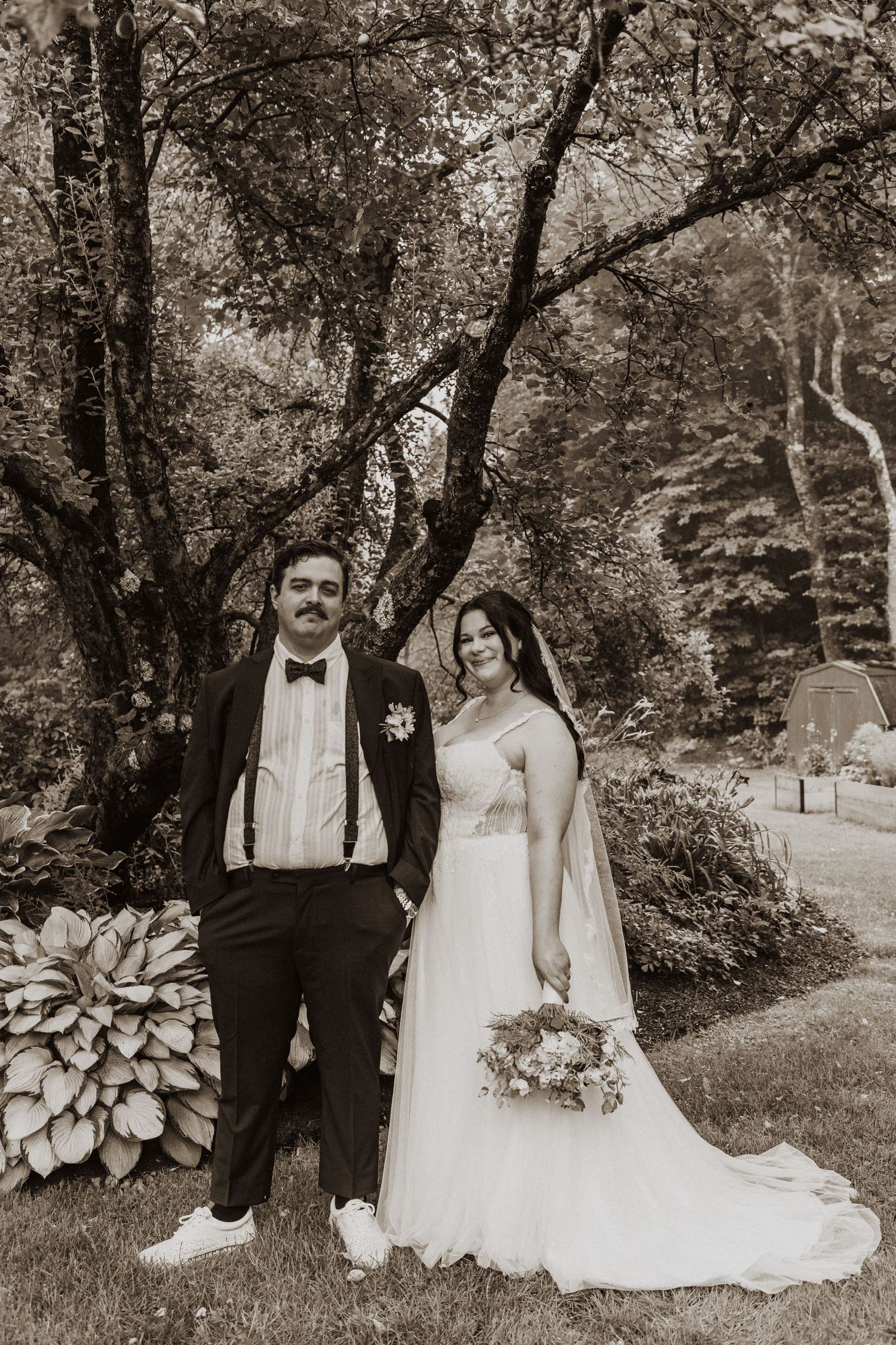 Sepia-toned wedding portrait of bride holding bouquet and groom in tuxedo standing in lush garden.