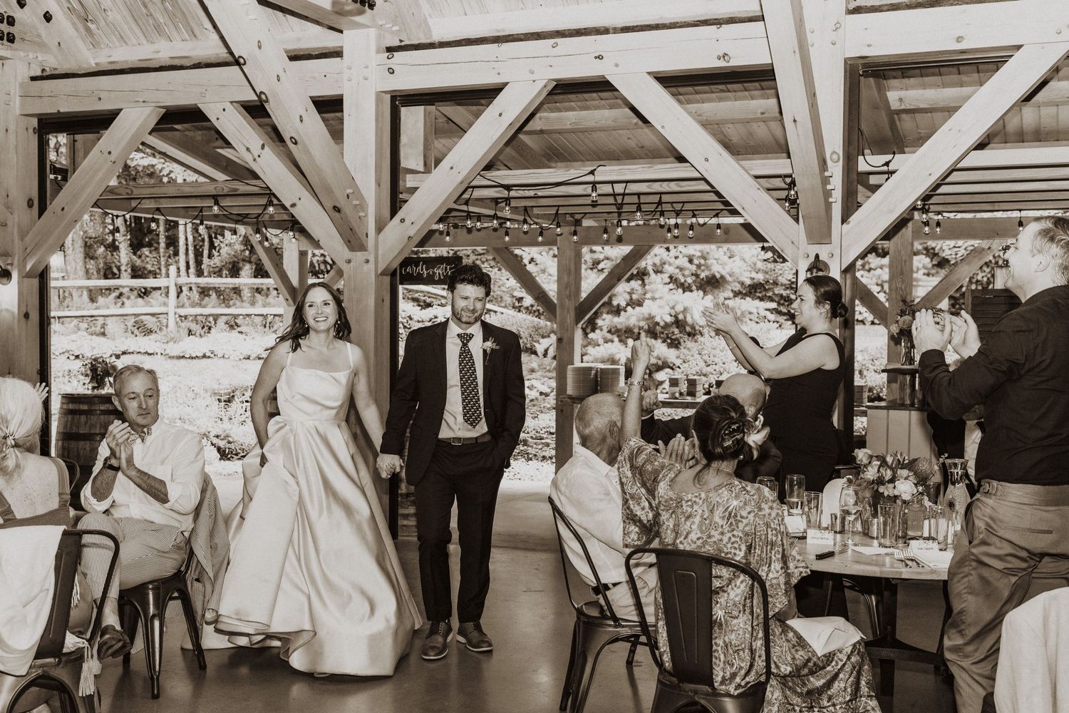 Black and white photo of bride and groom entering rustic outdoor wedding reception venue with seated guests.