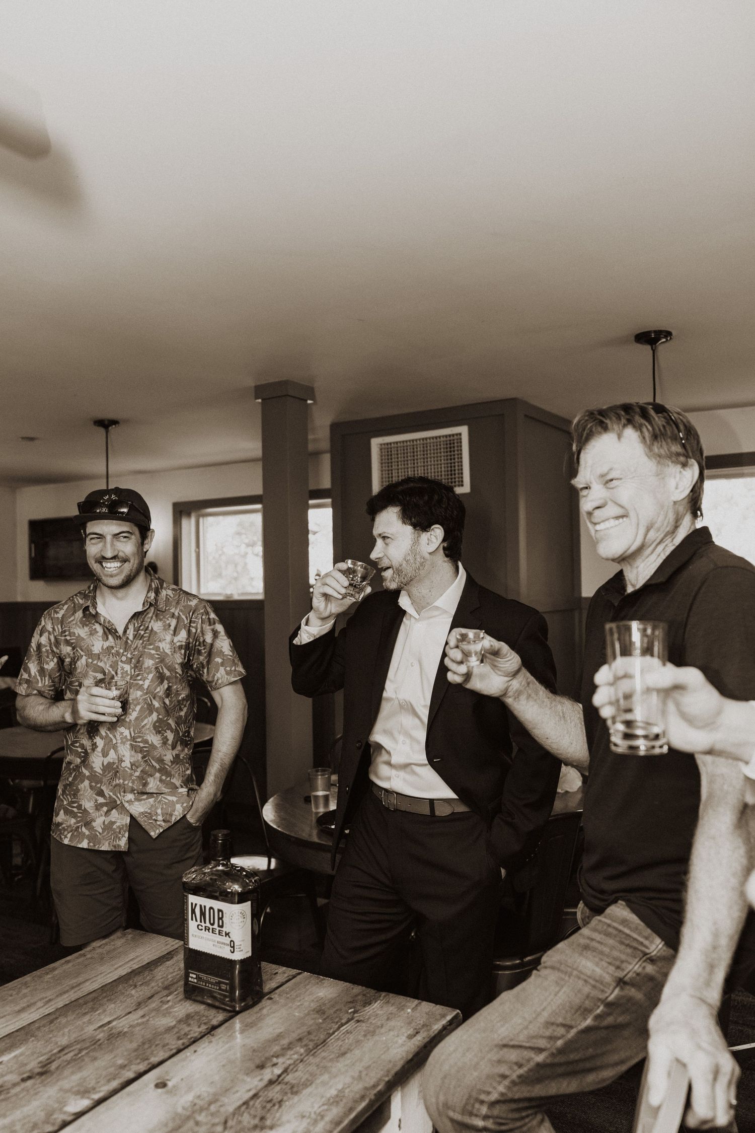Three men laughing and drinking whiskey shots together at a rustic wooden bar during a wedding celebration.