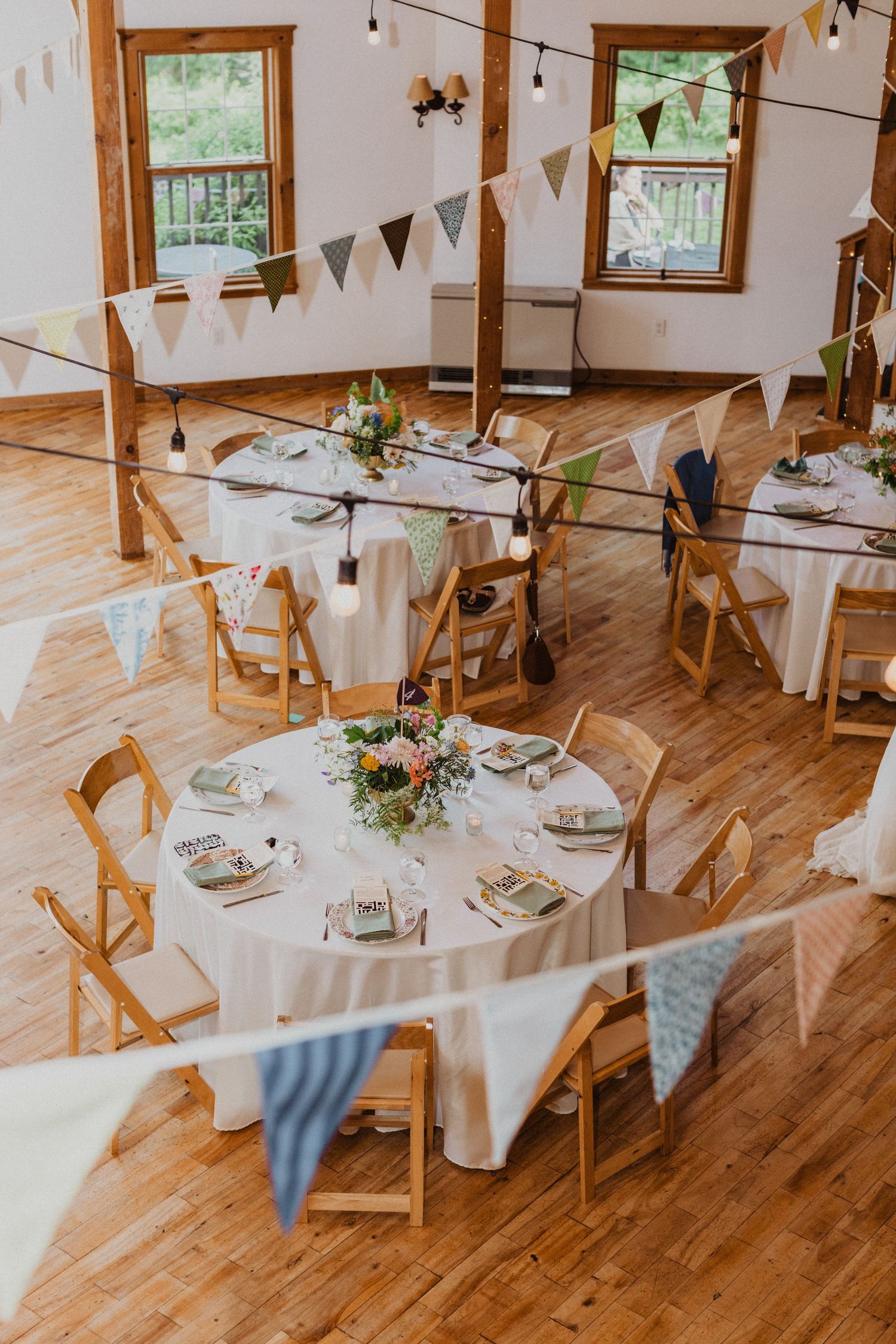 Rustic wedding reception hall with round tables, wooden chairs, and colorful bunting flags overhead.