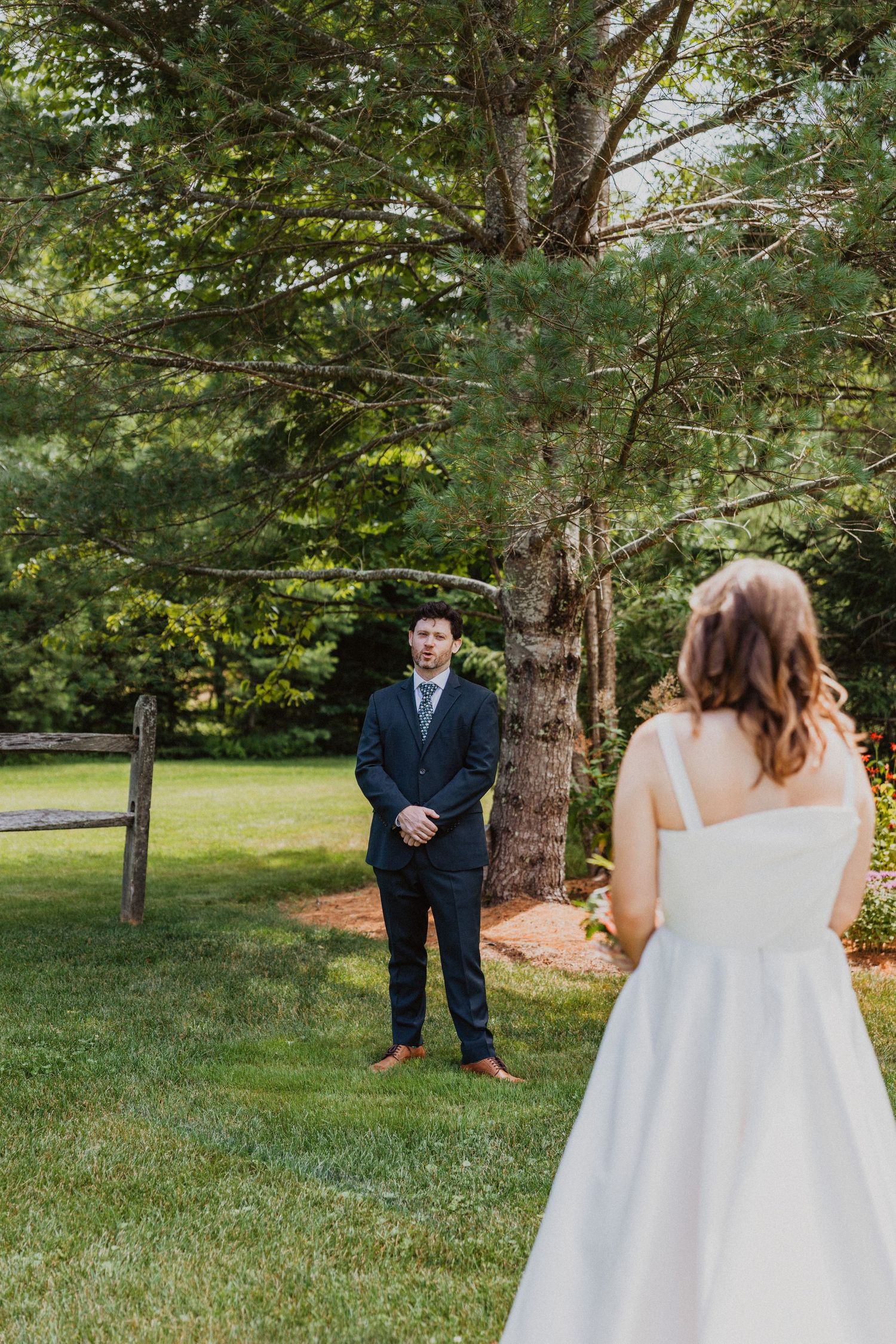 Groom in navy suit watching bride approach in white gown for romantic first look in green garden.