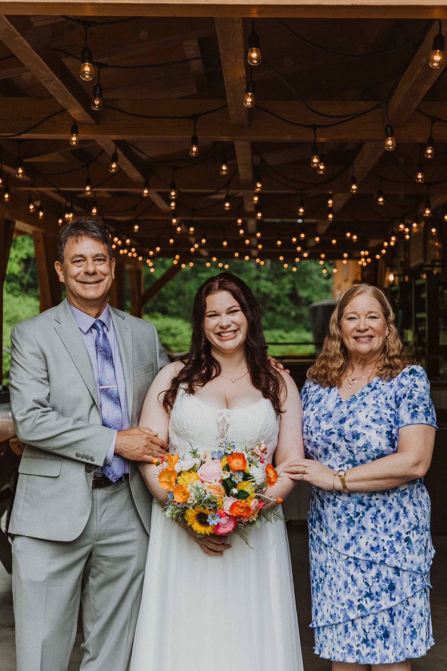 Smiling bride holding colorful bouquet poses with parents under string lights at rustic outdoor wedding venue.