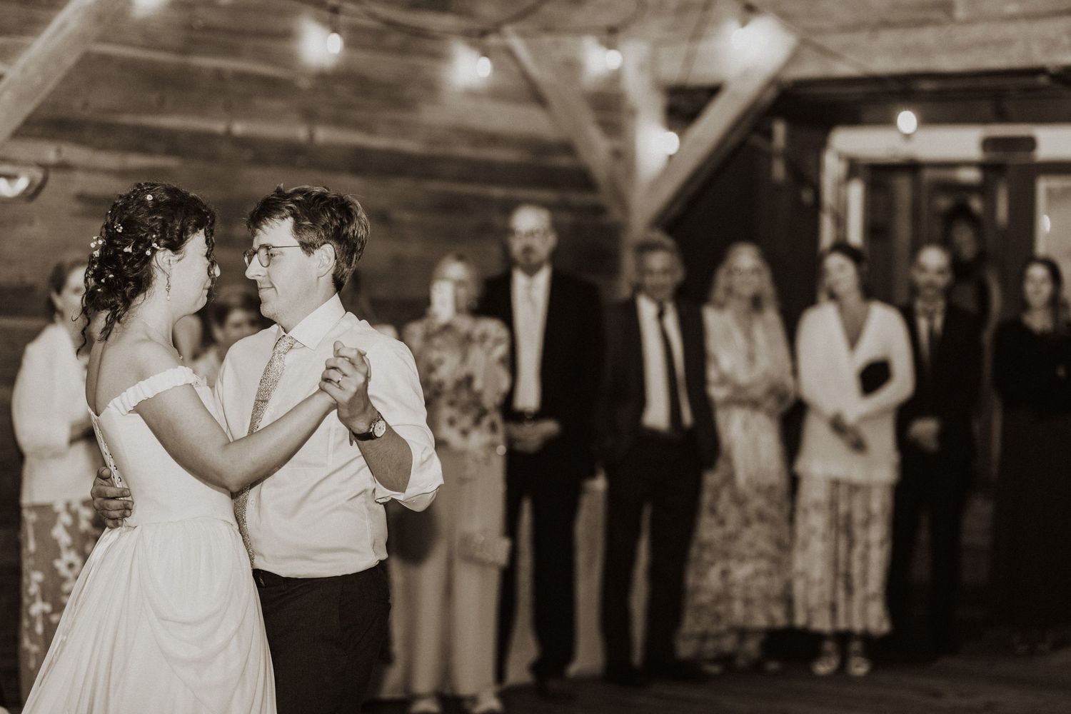 Bride and groom share first dance in rustic barn venue while wedding guests watch in background.