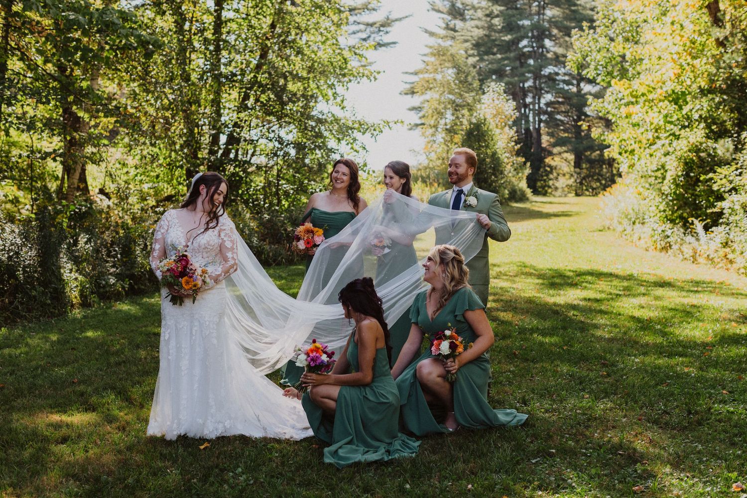 Bride in lace gown with long veil surrounded by bridesmaids in sage green dresses at outdoor wedding.