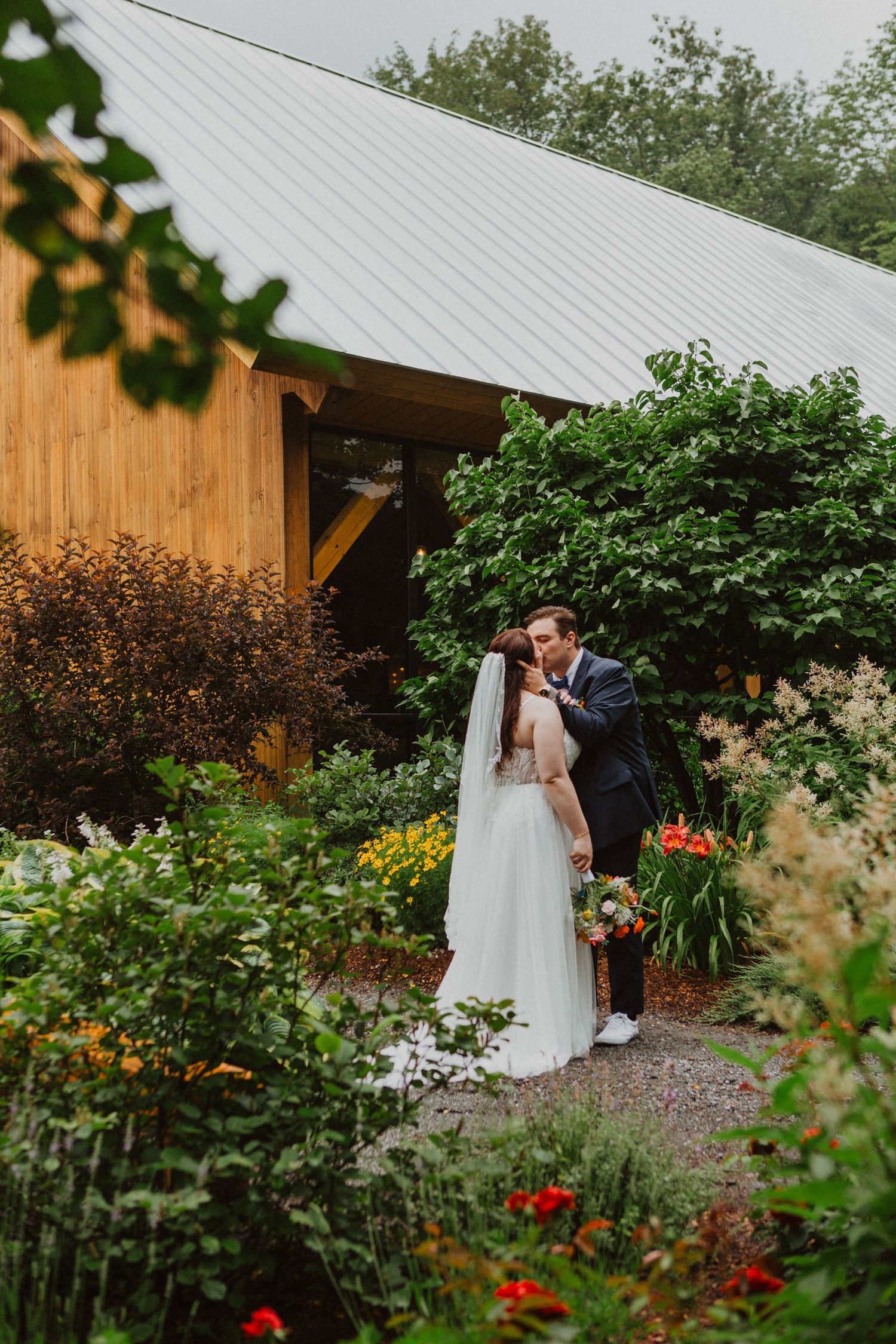 Bride and groom sharing a kiss in a lush garden outside a rustic wooden wedding venue.