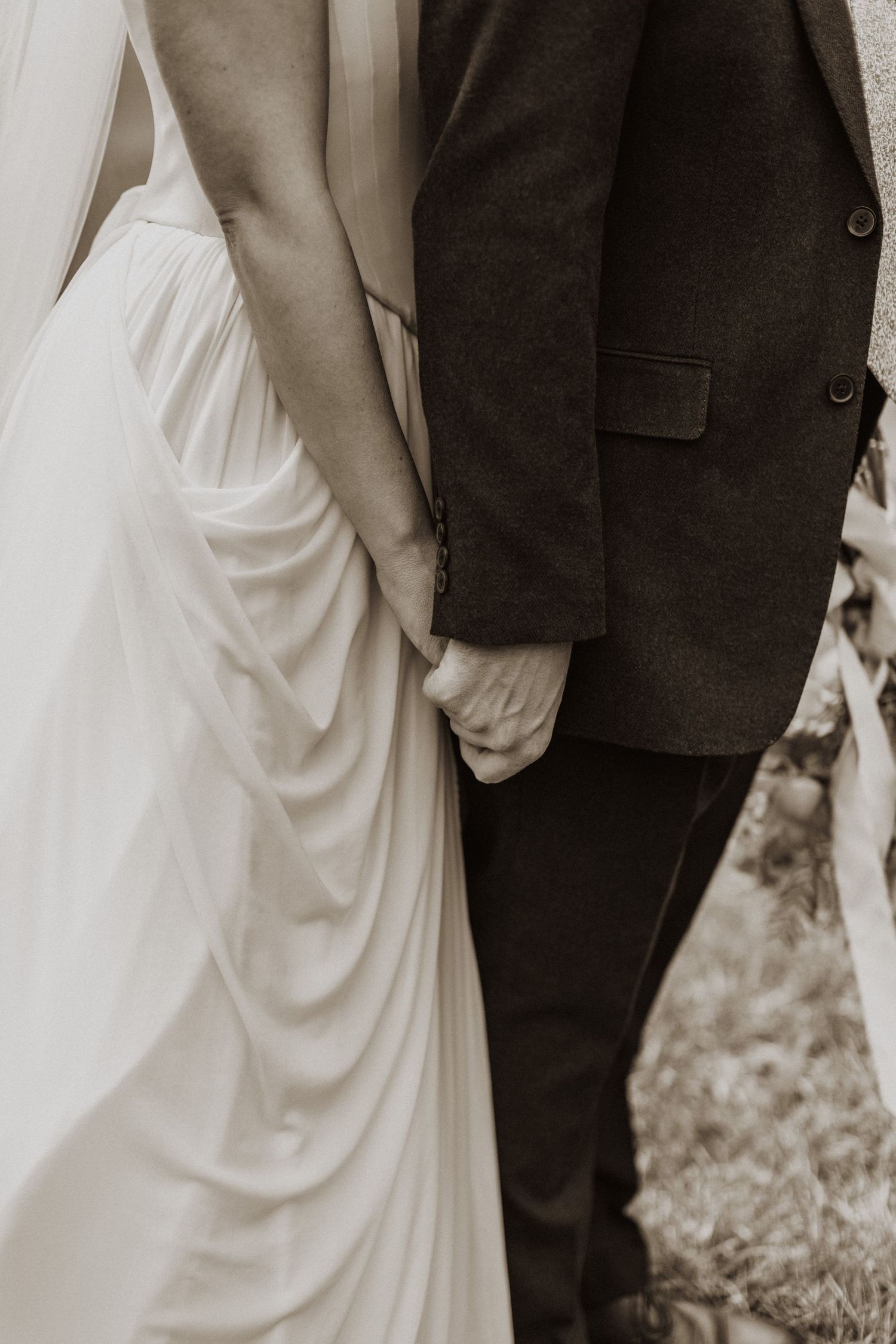 Close-up of bride and groom holding hands, wedding dress and suit detail, sepia tone.