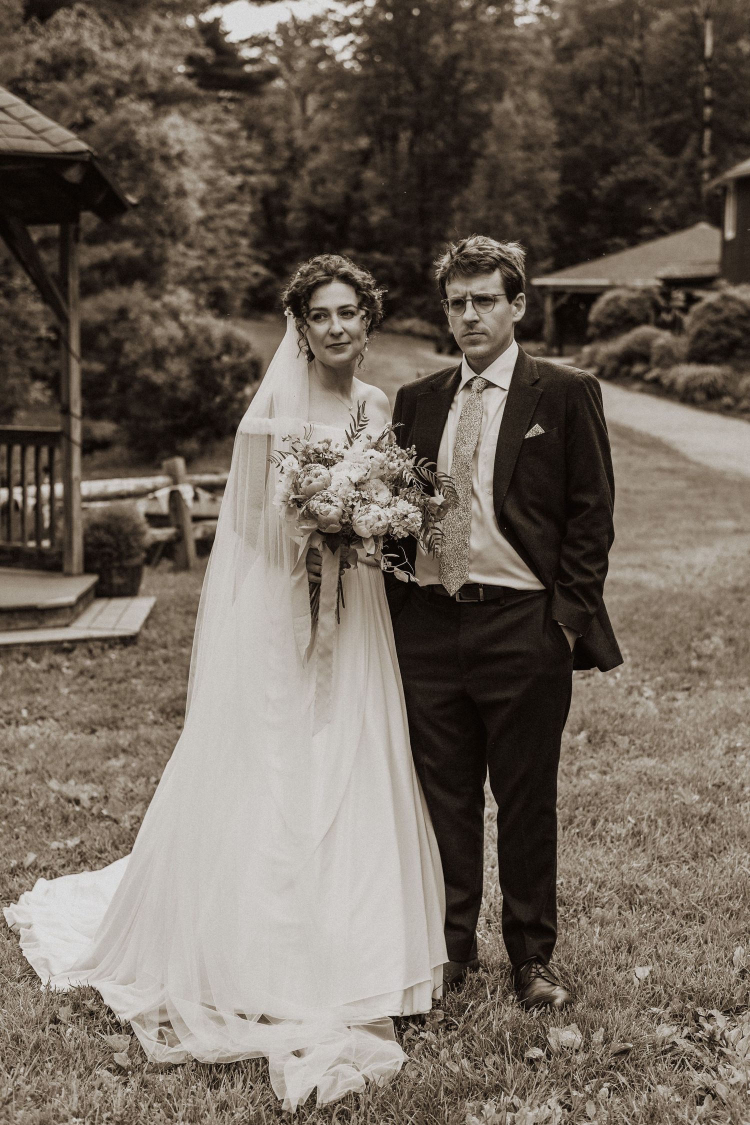 Sepia toned portrait of bride holding bouquet and groom standing together on lush green lawn.