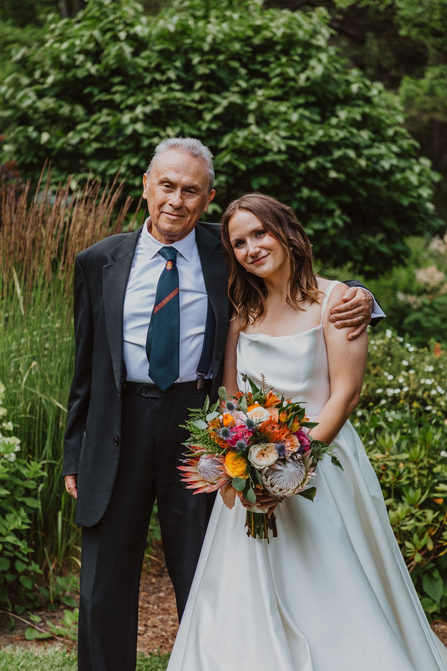 Bride in white gown holds colorful bouquet beside elderly man in dark suit outdoors.