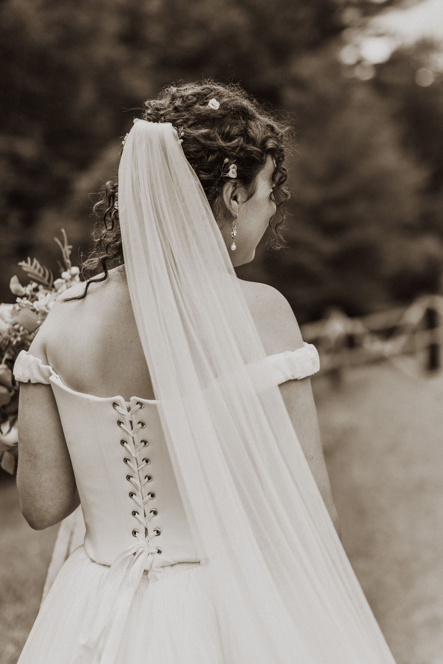 Bride in off-shoulder lace-up wedding gown with long veil, sepia-toned back view.
