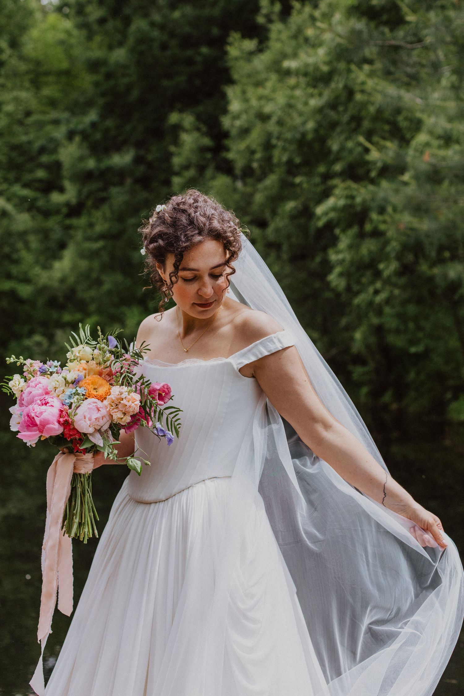 Bride in white off-shoulder gown holding colorful bouquet with veil flowing outdoors near greenery.