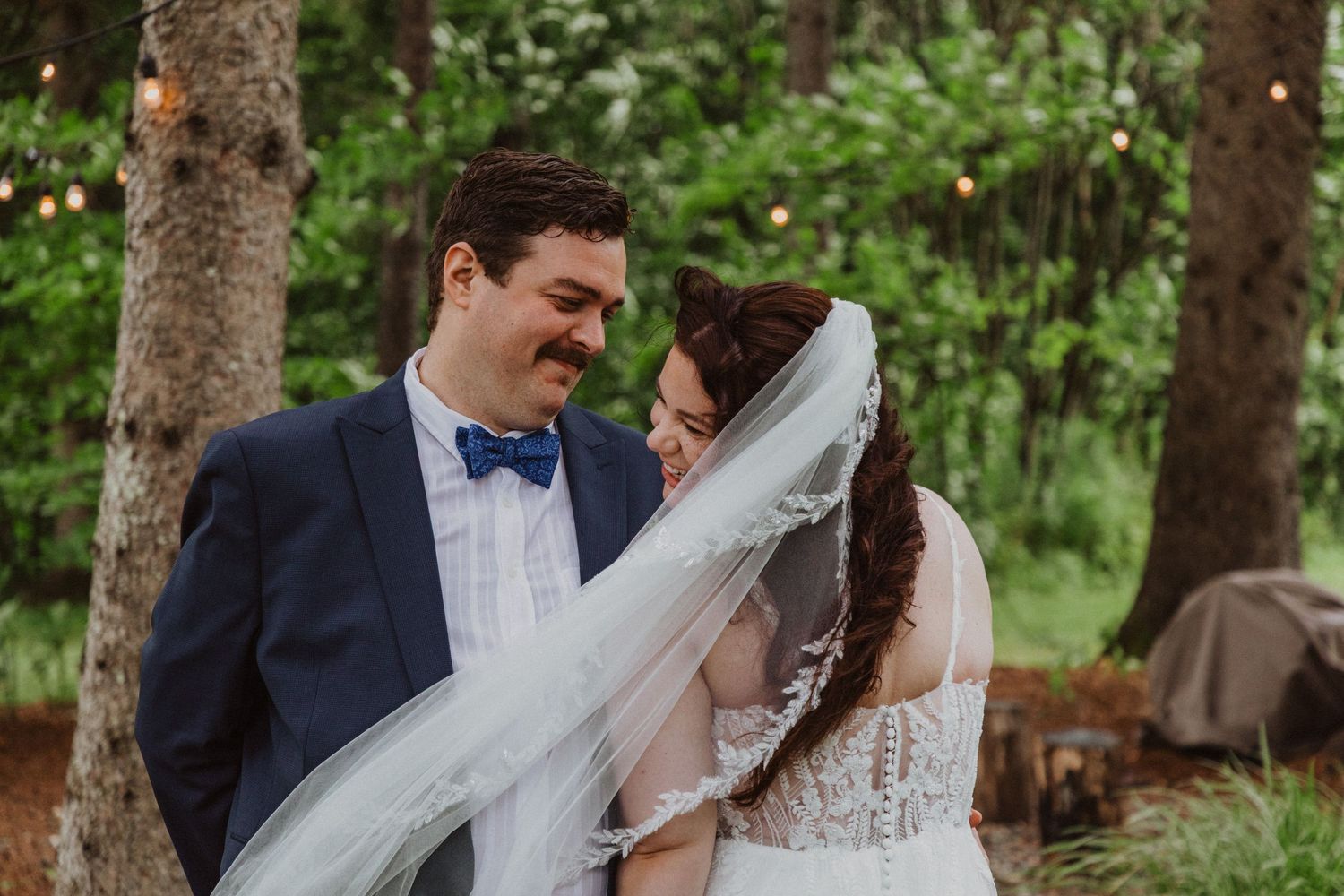 Bride and groom share an intimate moment among pine trees, her veil flowing softly in a lush forest setting.
