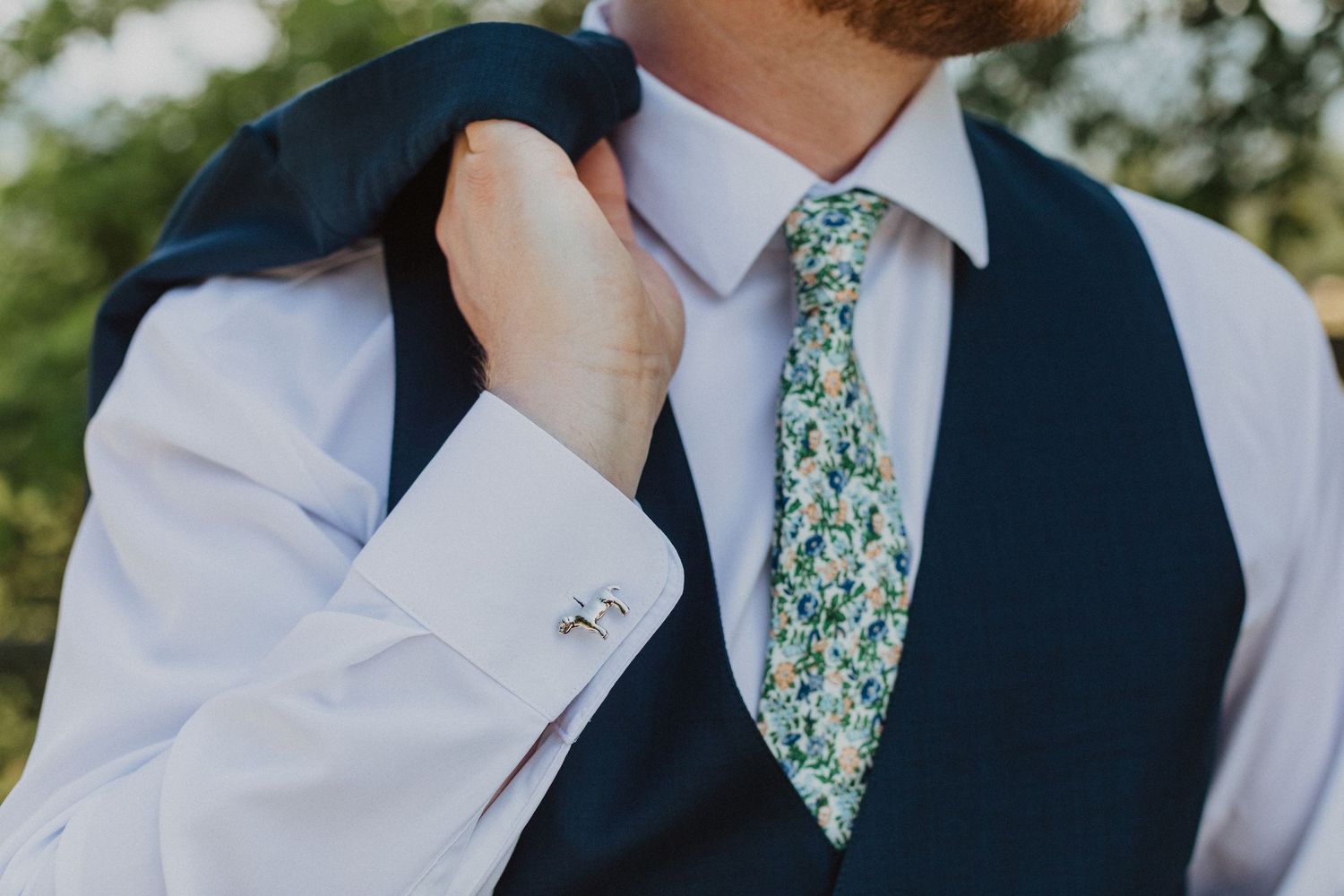 Close-up of groom in navy vest with floral green tie and silver cufflinks on wedding day.
