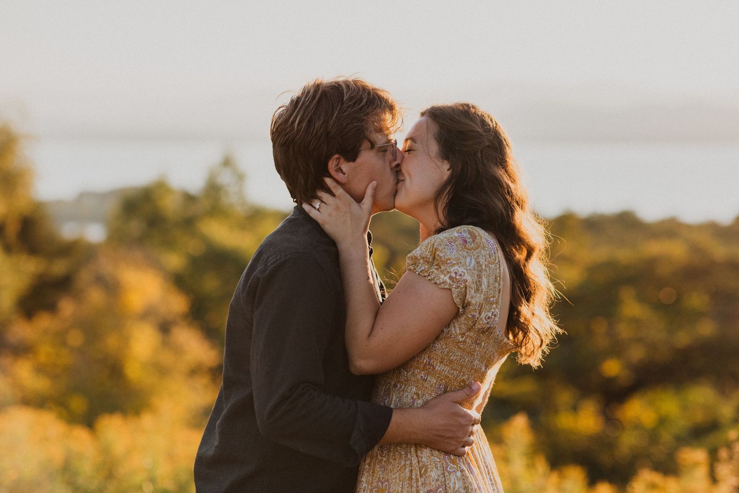 A couple shares a tender kiss at golden hour surrounded by vibrant autumn foliage and a scenic lake view.