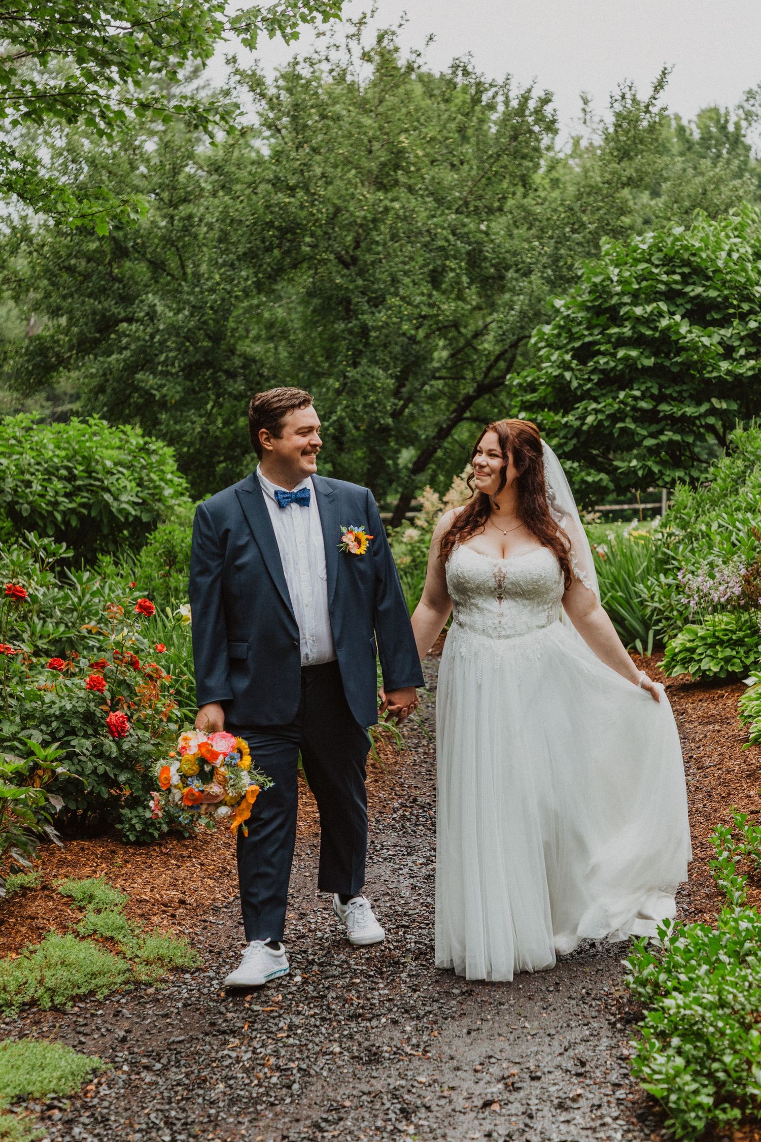 Bride and groom walk hand-in-hand through a lush garden path, smiling at each other, holding a colorful bouquet.