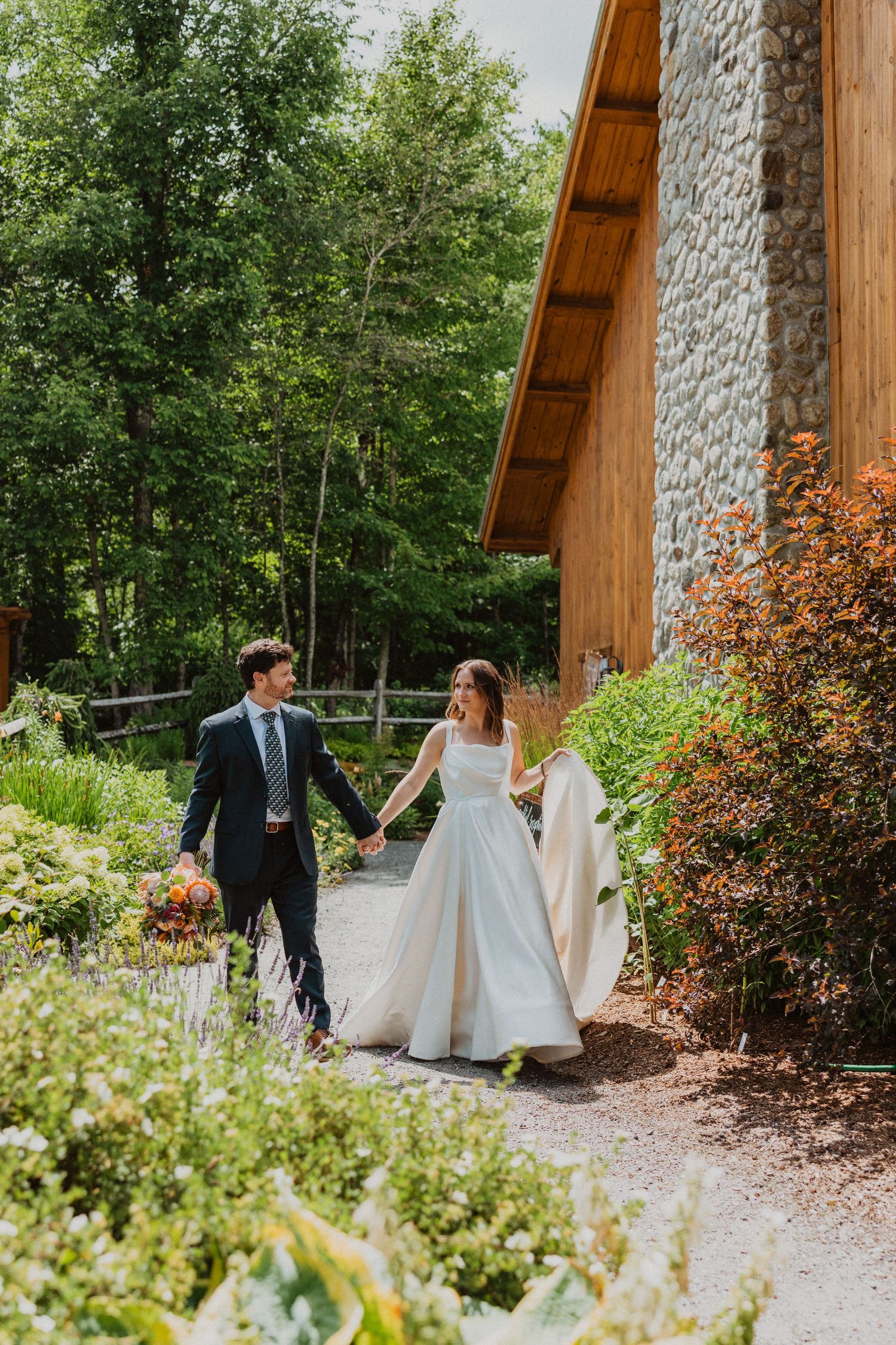 A bride and groom walk hand-in-hand along a garden path beside a rustic stone and wood building surrounded by lush greenery.
