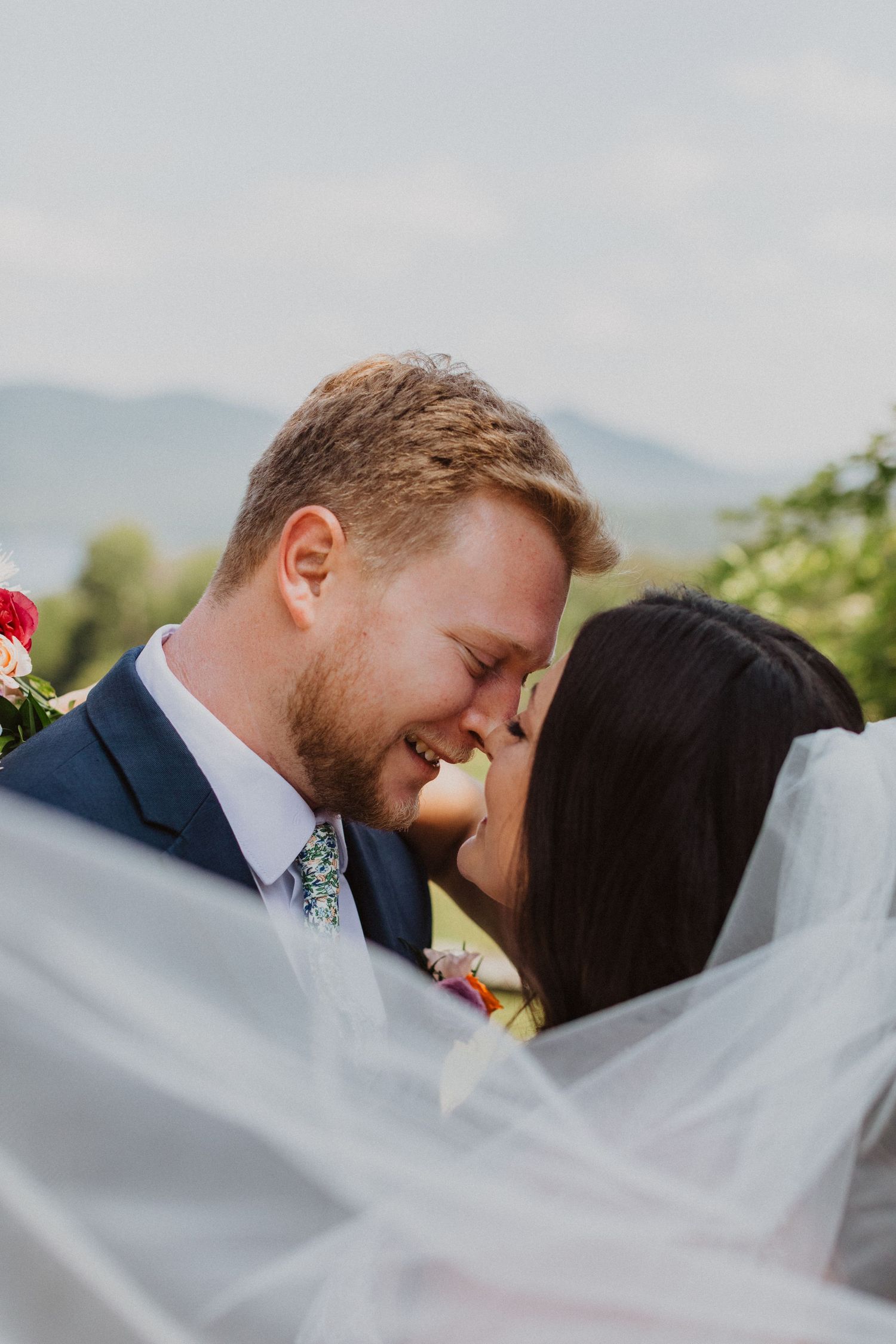 Smiling bride and groom nearly kissing under flowing veil with mountain backdrop on wedding day.
