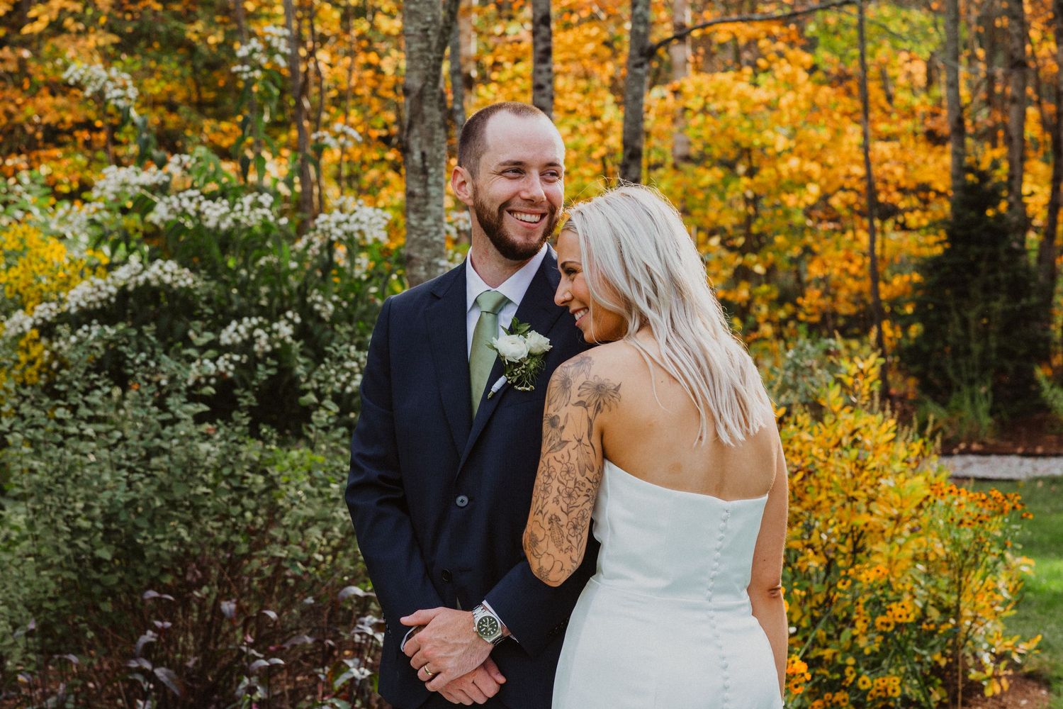 A smiling groom in a navy suit embraces his blonde bride in a strapless gown amid colorful fall foliage.