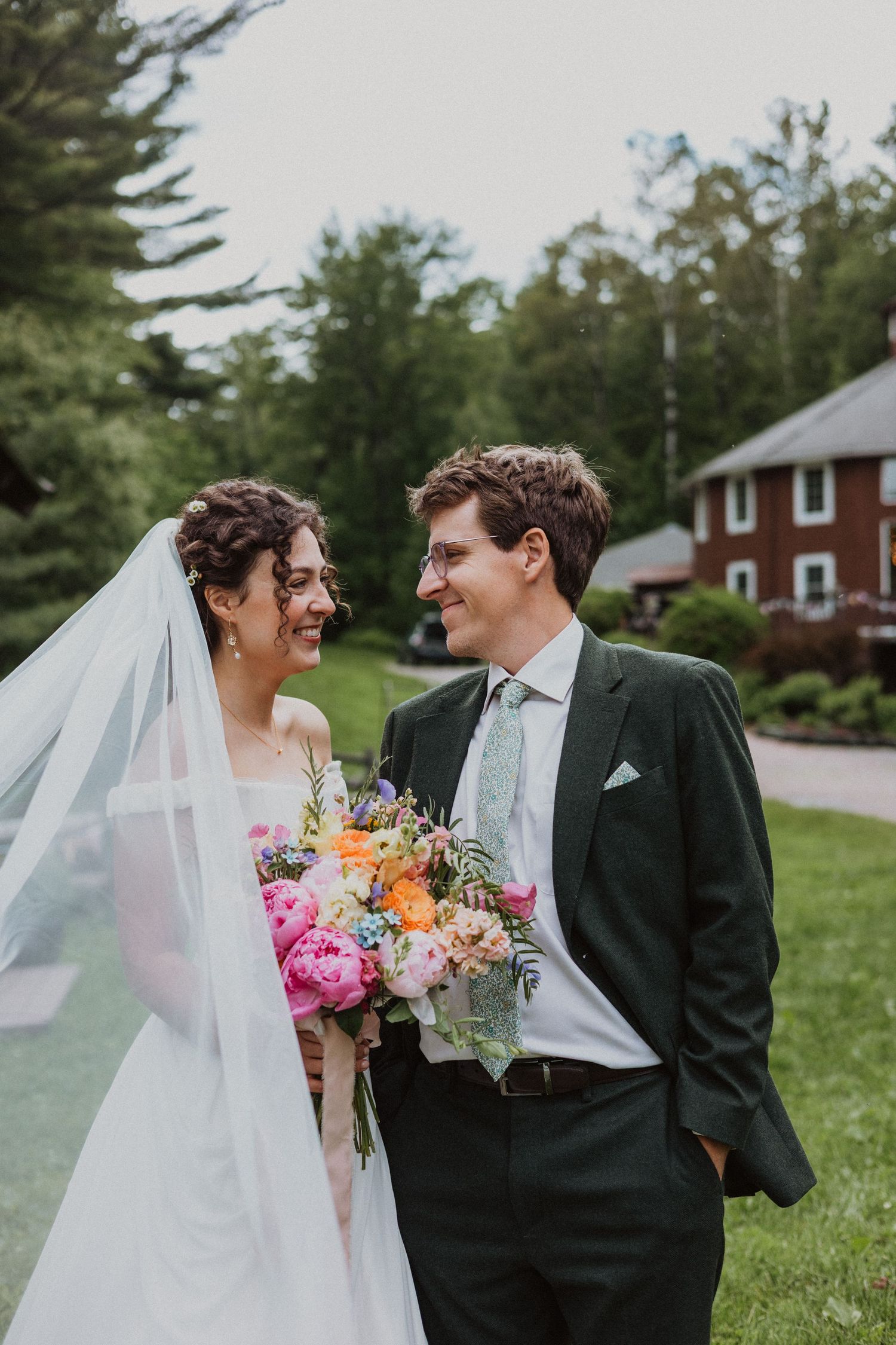 Bride and groom smiling at each other outdoors, bride holding colorful bouquet with veil flowing, lush green trees behind.