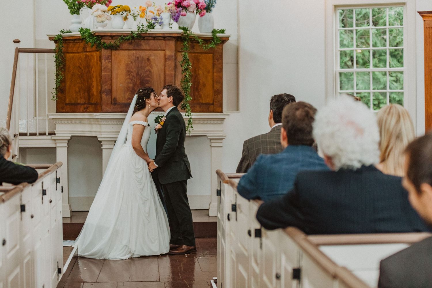 Bride and groom sharing first kiss at altar in a white chapel with guests seated in wooden pews.
