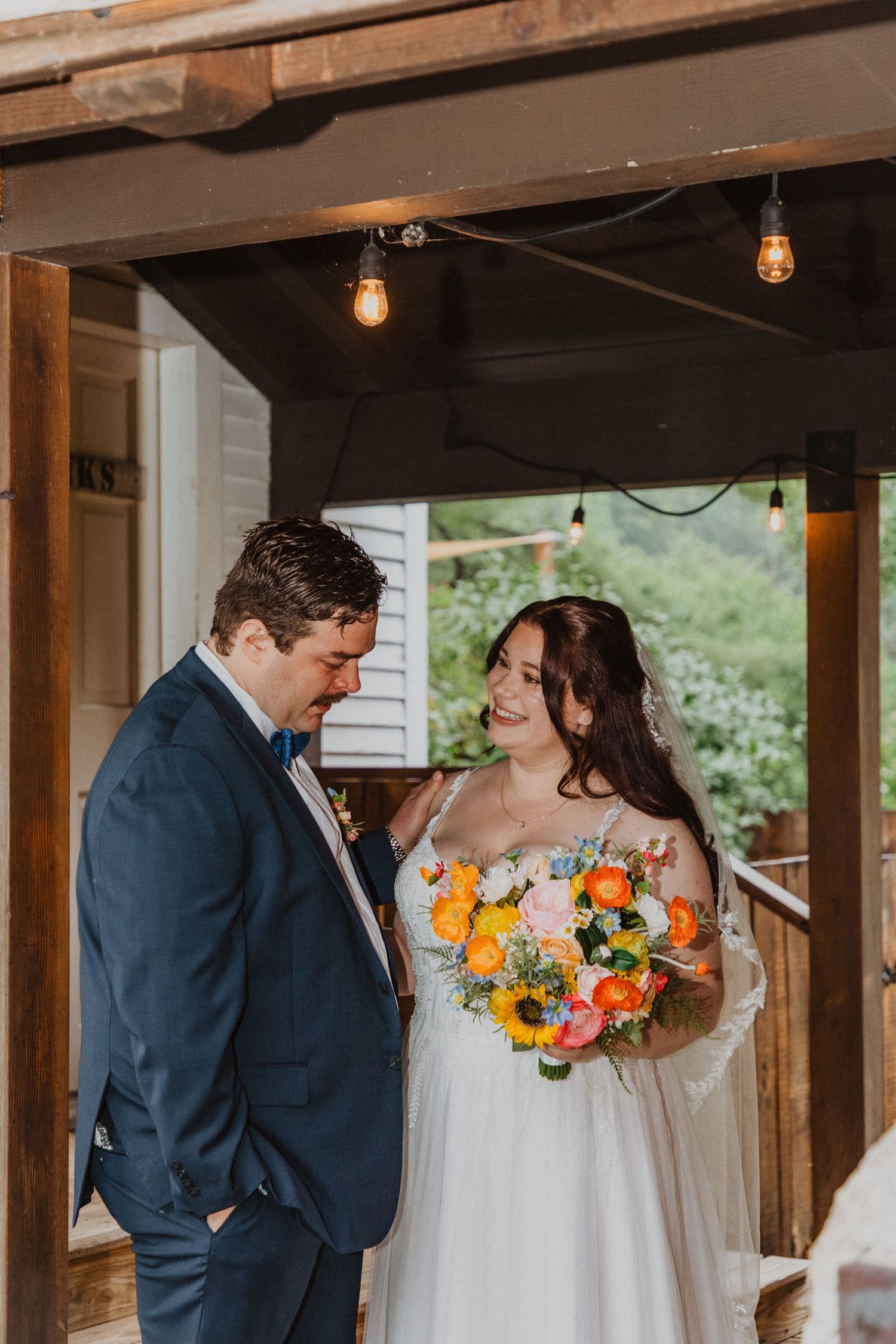 Bride and groom share a tender moment under a rustic wooden pergola with string lights, bride holding a vibrant bouquet.
