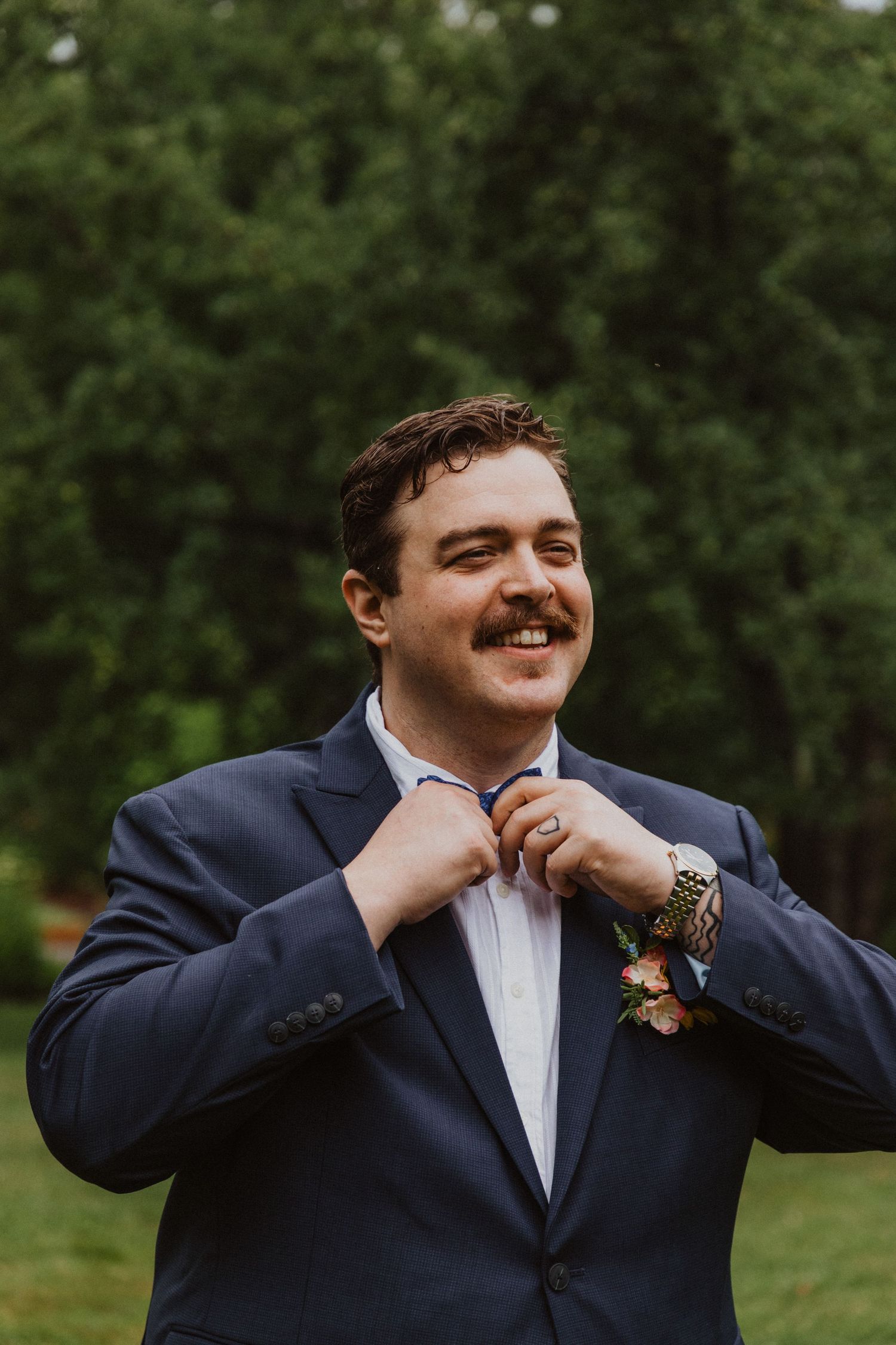 Smiling groom in navy suit adjusting bow tie with boutonniere and decorative watch outdoors.