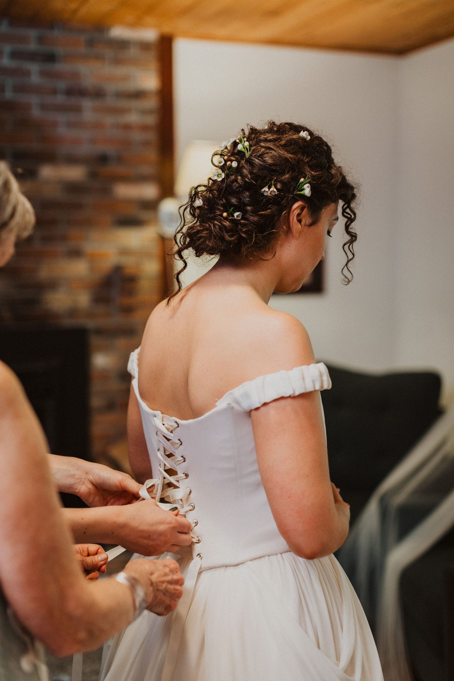 Bride in off-shoulder corset wedding gown getting laced up by assistant in rustic wood-paneled room.
