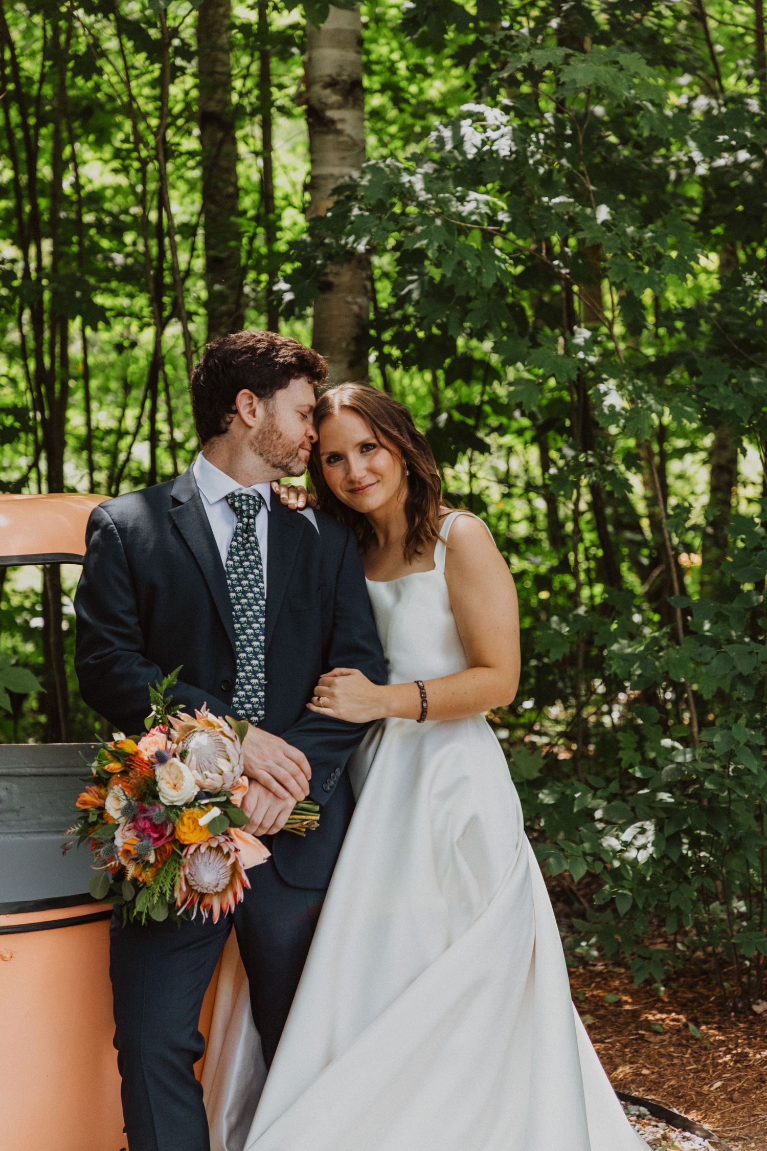 Bride and groom posing in lush forest setting, groom in navy suit with green tie holding colorful wedding bouquet.