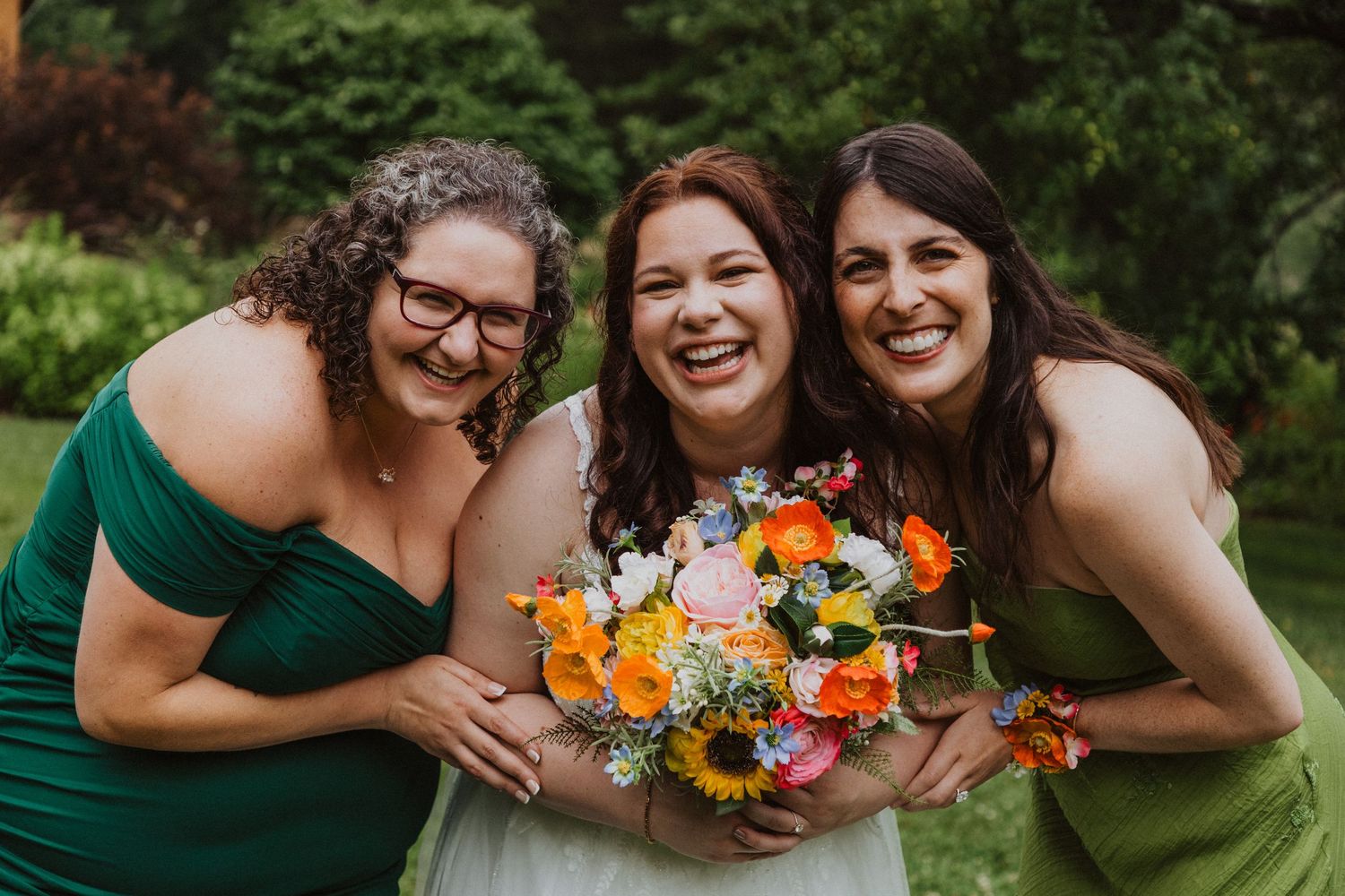 Three joyful women smile together outdoors, bride holding vibrant orange and yellow bouquet flanked by bridesmaids in green.