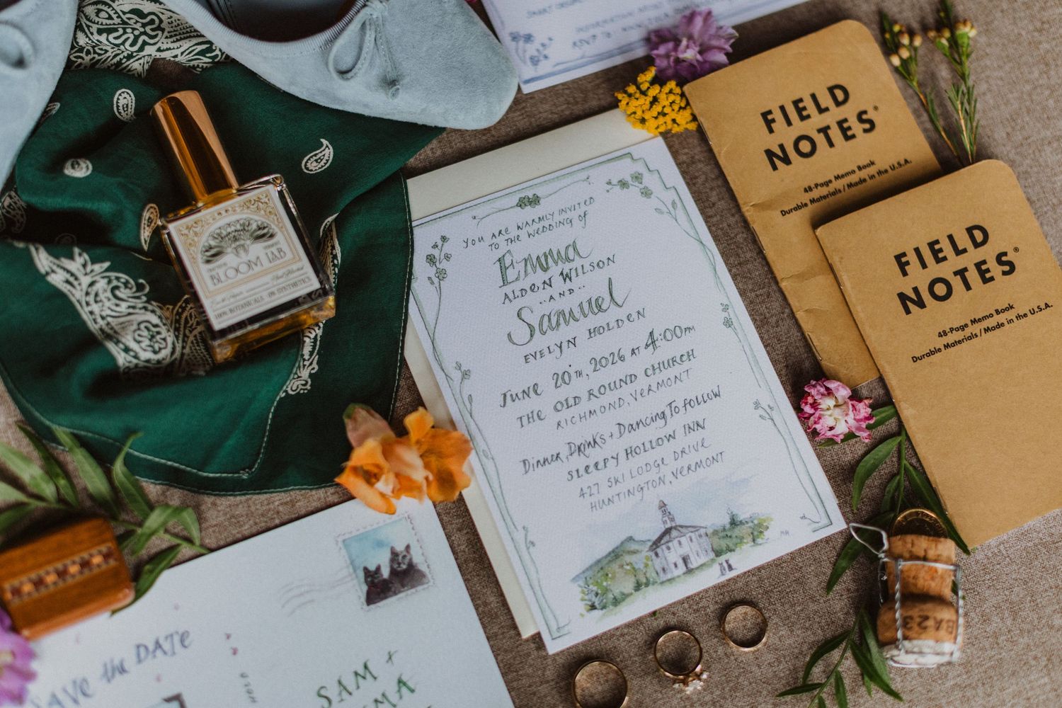 Rustic wedding stationery flat lay featuring invitations, Field Notes notebooks, rings, and perfume on a textured surface.