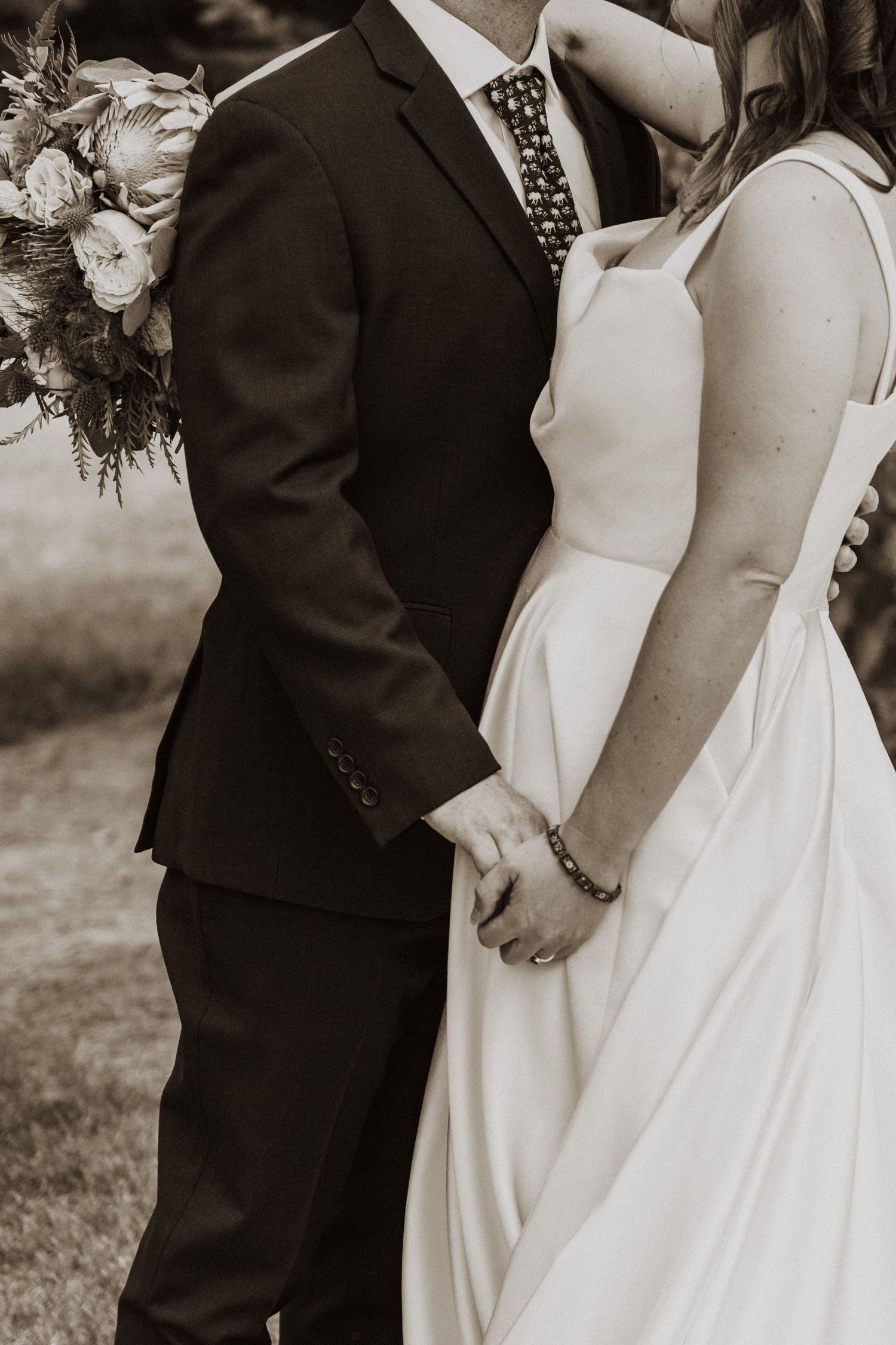 Black and white photo of bride and groom holding hands, bride in white gown, groom in dark suit with floral bouquet.