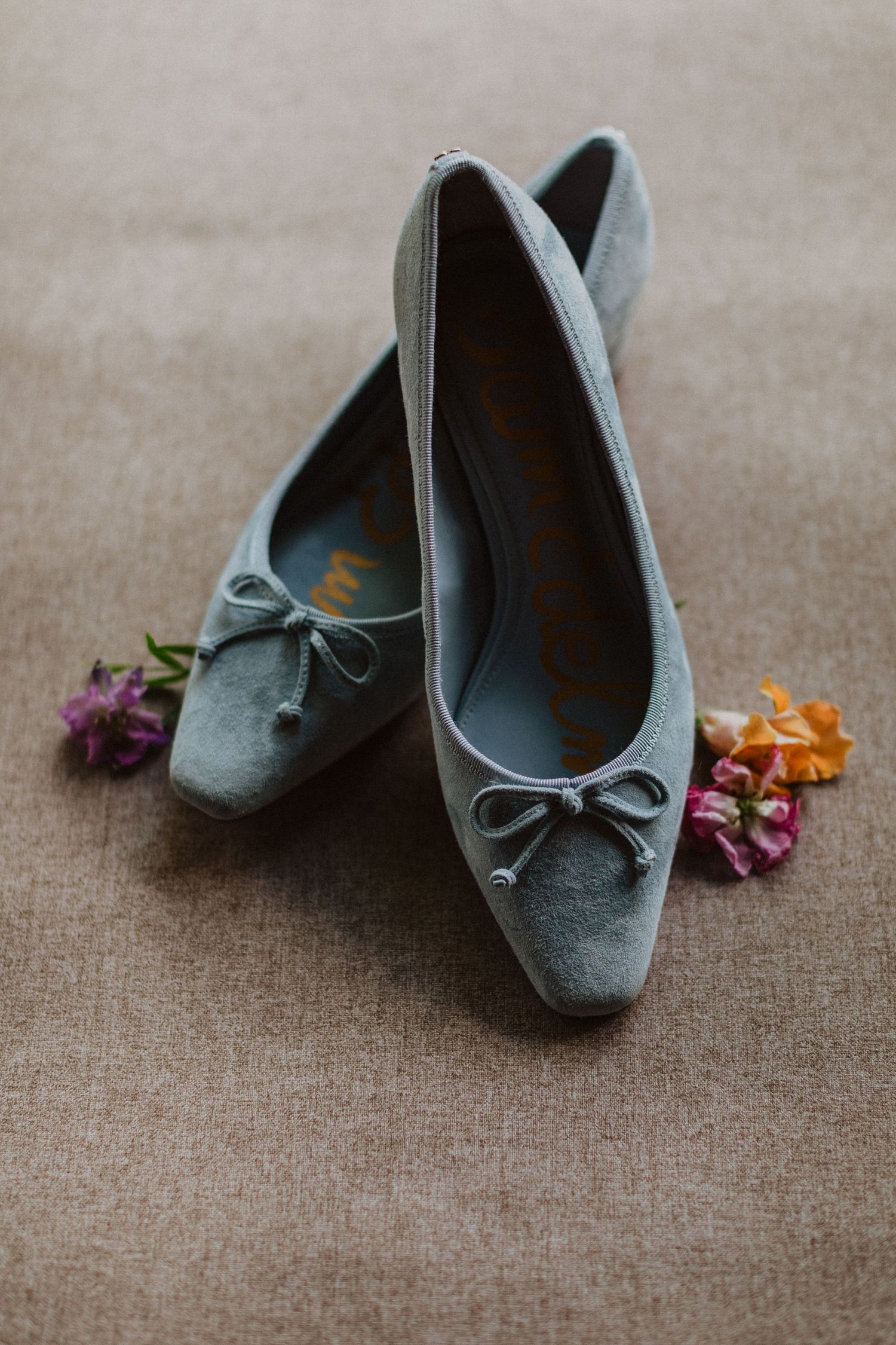 Light blue suede ballet flats with bow detail surrounded by colorful wildflowers on linen.