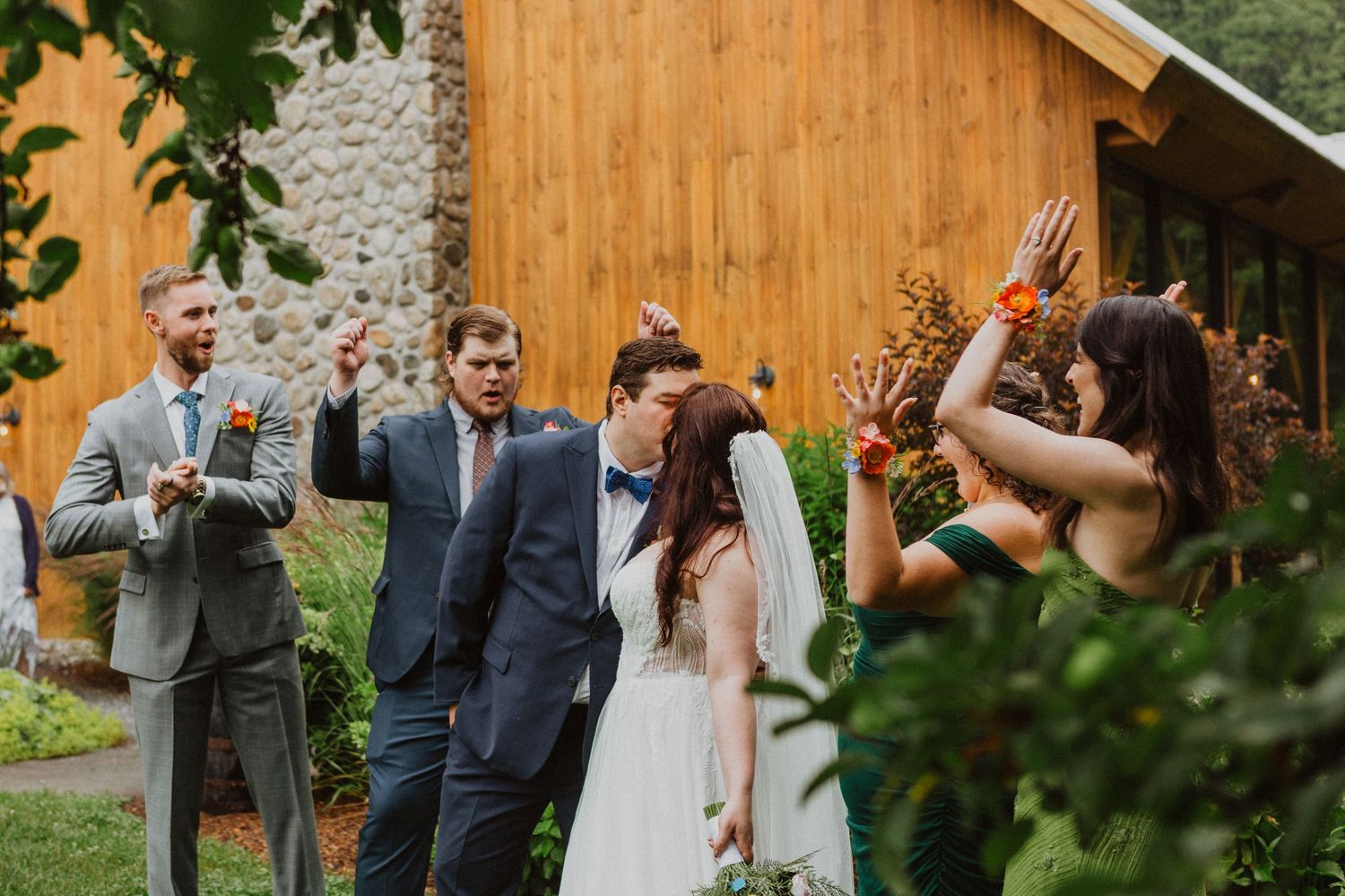 Wedding guests celebrate outdoors near a wooden barn as a bride and groom share a kiss during their ceremony.