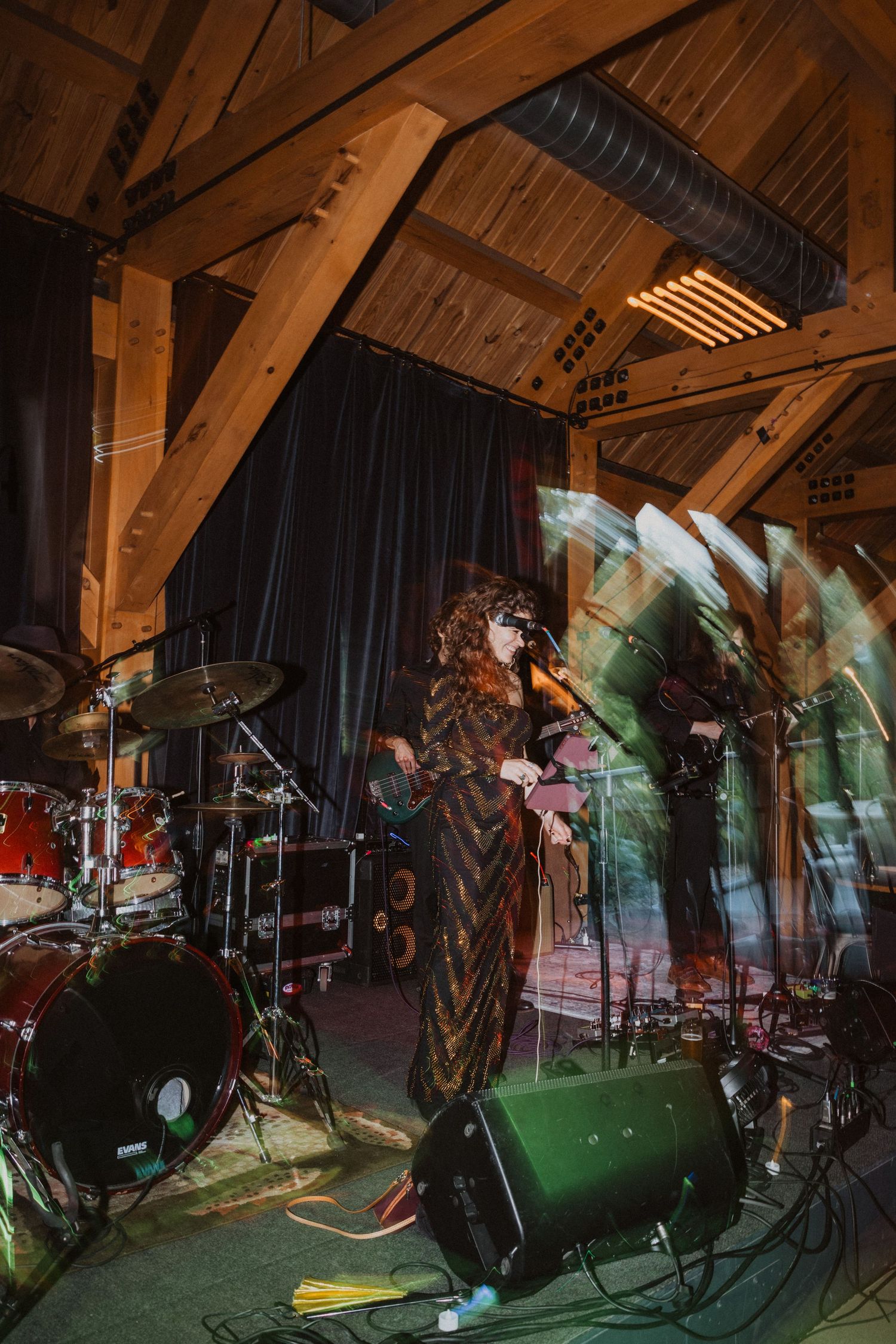 Live band performing on stage in a rustic wooden venue with dramatic lighting and a curly-haired singer in a patterned dress.