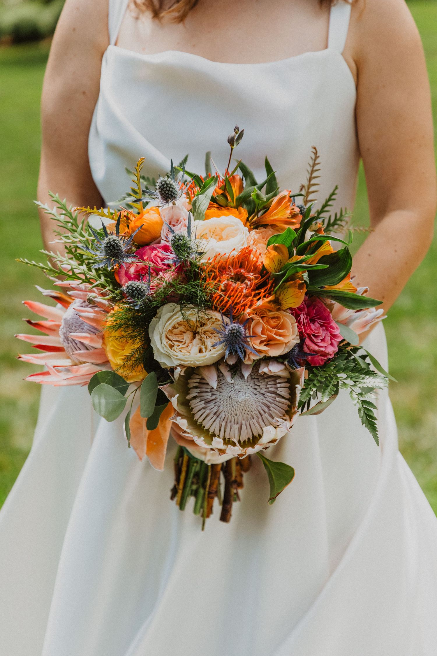 Bride holding vibrant wedding bouquet with protea, roses, and thistle in white gown outdoors.