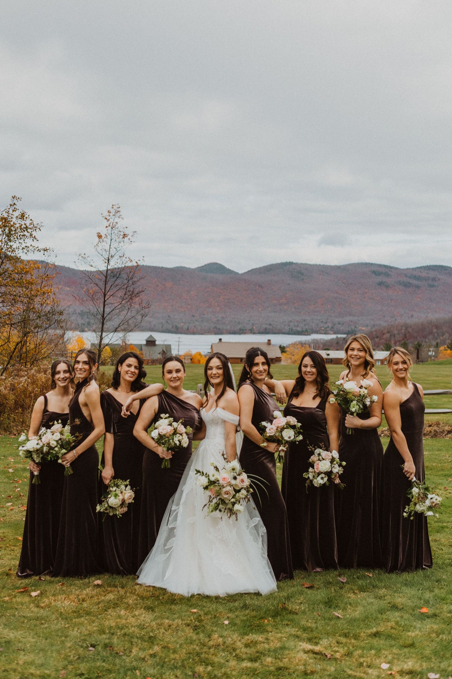 Bride in white gown with eight bridesmaids in dark velvet dresses holding bouquets in mountain setting.