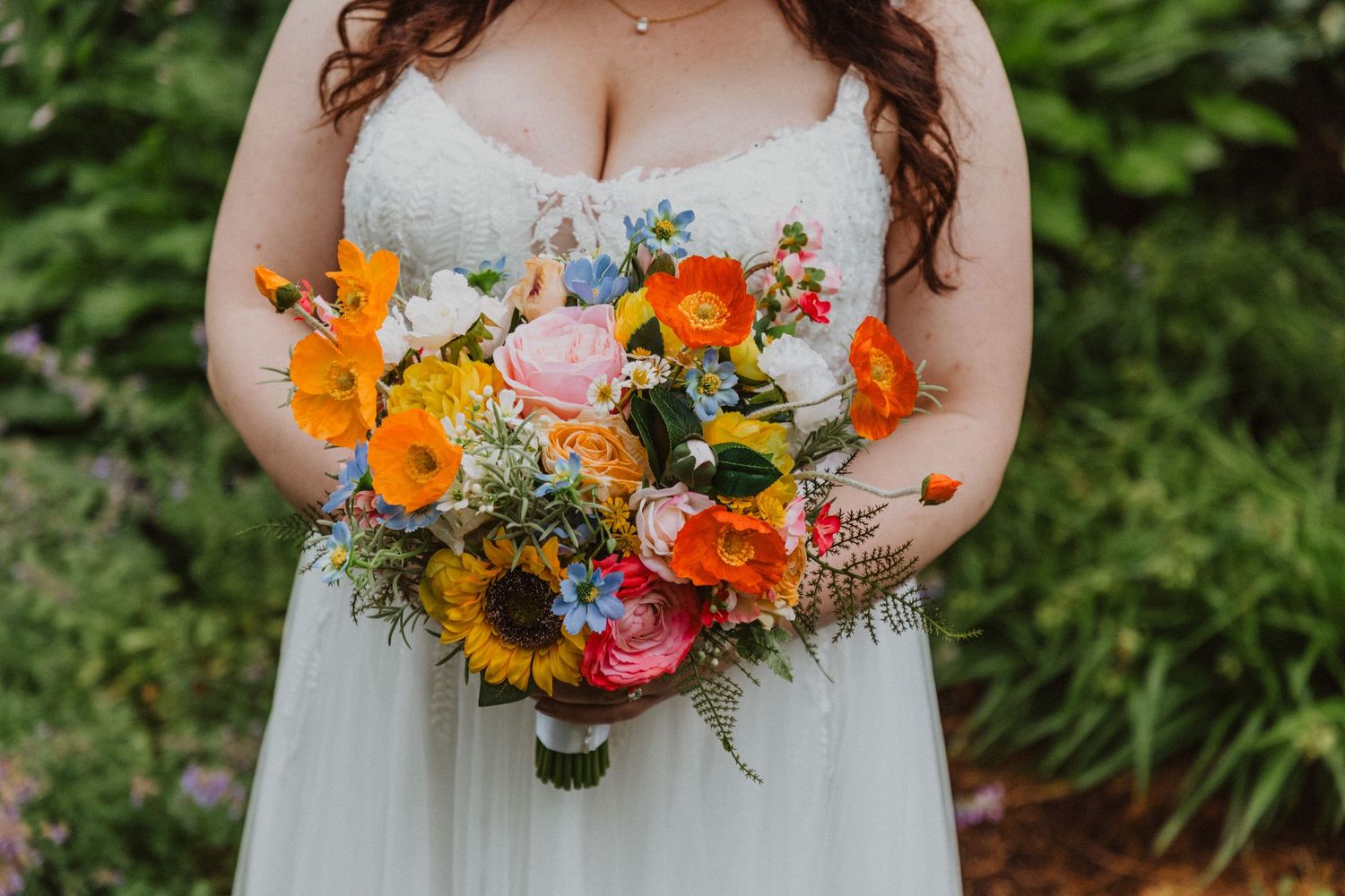 Bride holds a vibrant wedding bouquet featuring sunflowers, orange gerberas, pink roses, and blue wildflowers.