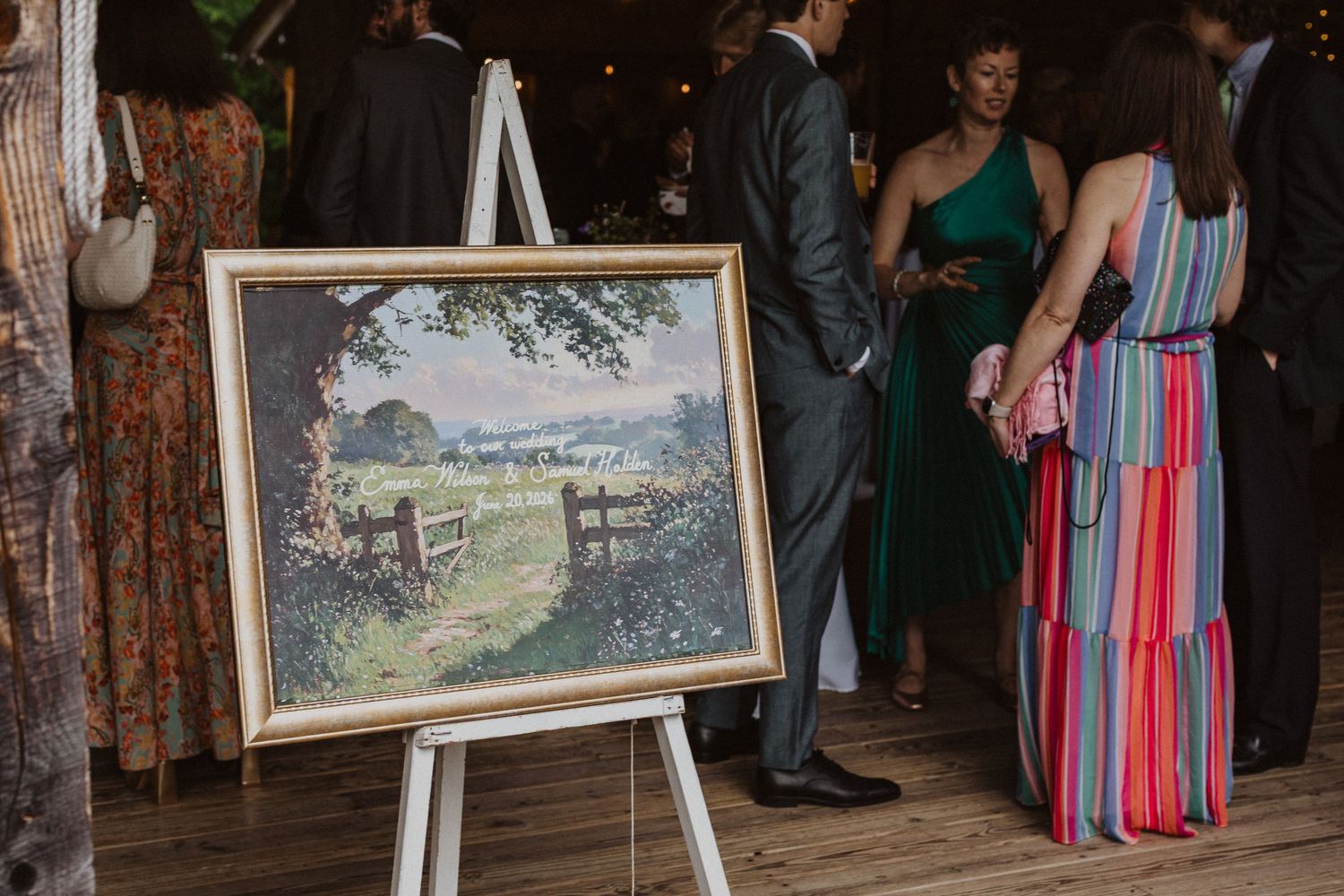 Wedding guests mingle near a framed landscape painting displayed on an easel at a barn reception.