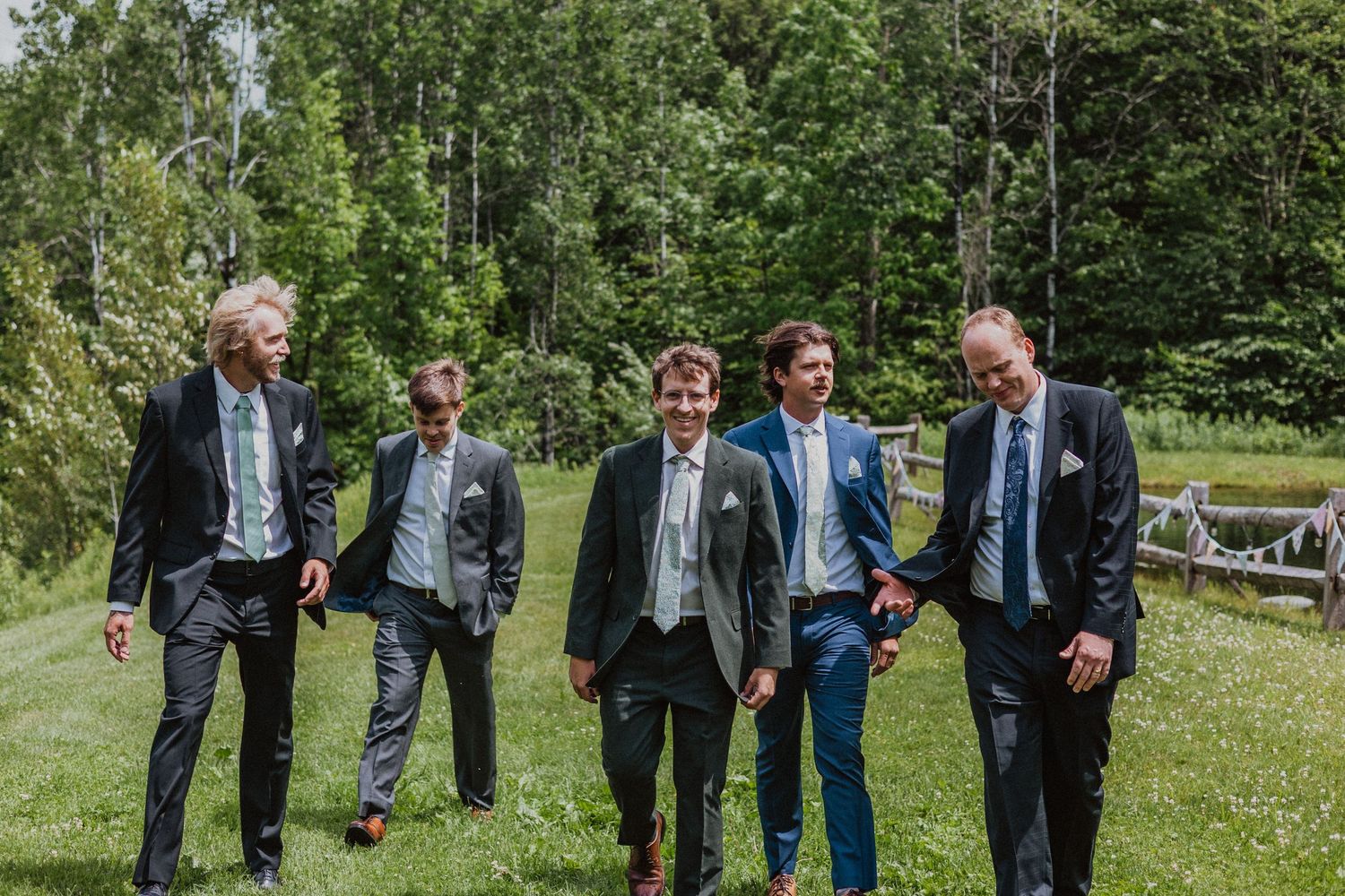 Five groomsmen in suits walking together on green grass at an outdoor wedding venue surrounded by trees.