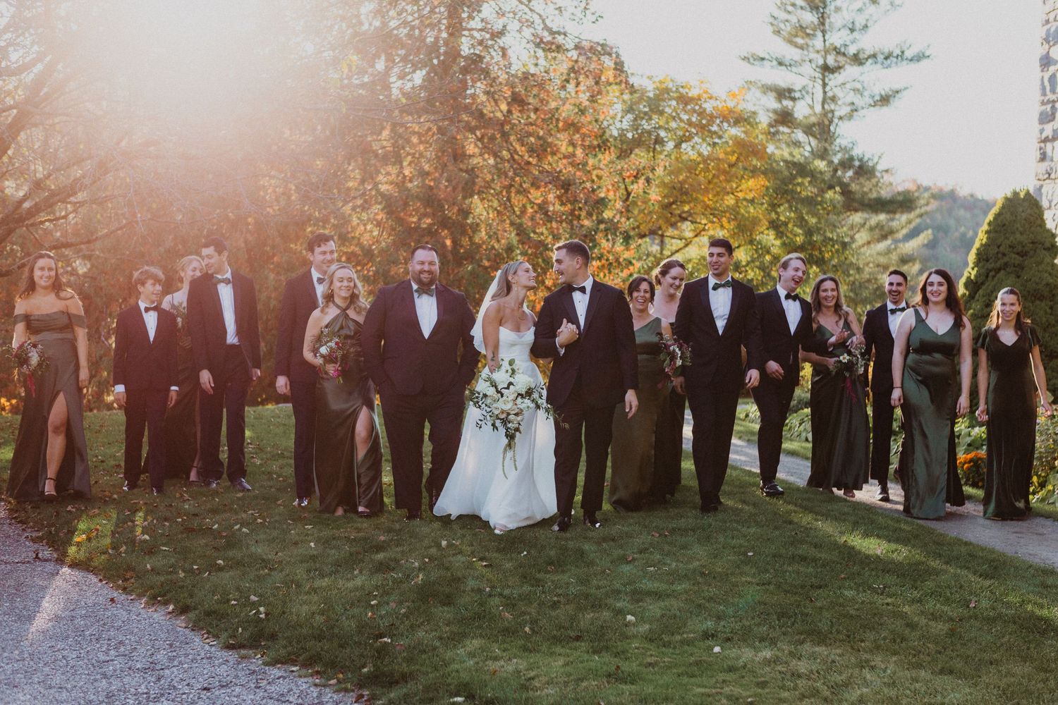 Wedding party walks together in golden autumn sunlight with bridesmaids in black and groomsmen in tuxedos.