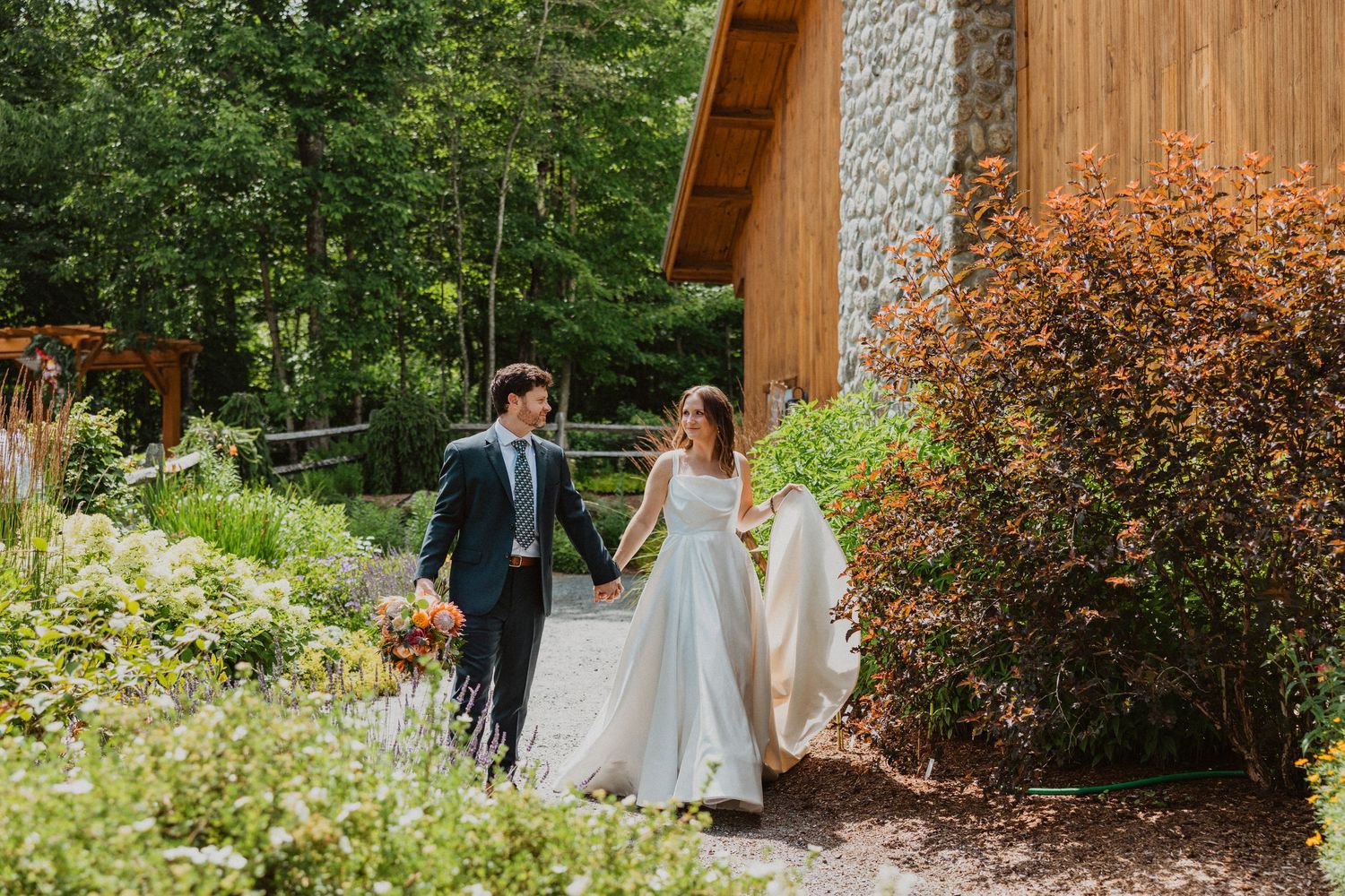 Bride and groom walk hand-in-hand through lush garden path beside rustic stone building on wedding day.