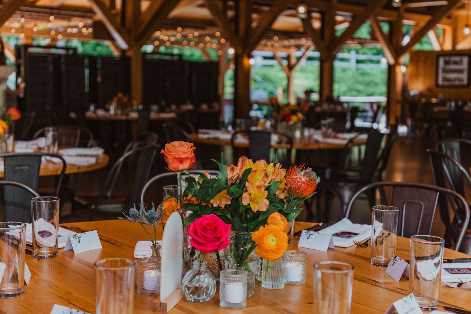 Rustic barn wedding reception with wooden tables, colorful floral centerpieces featuring pink, orange roses and alstroemeria blooms.