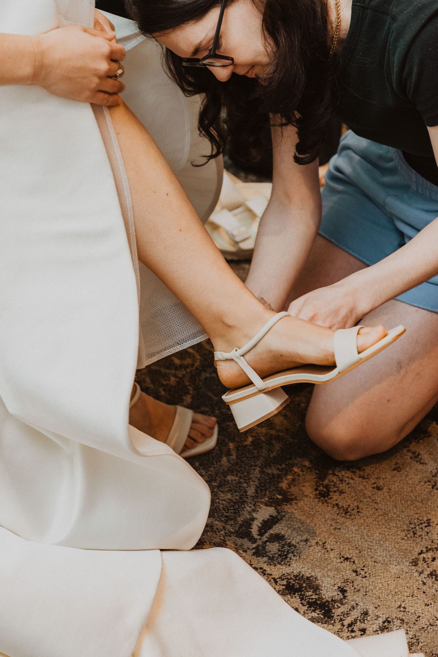 Bridesmaid helping bride put on white block heel sandals before wedding ceremony.