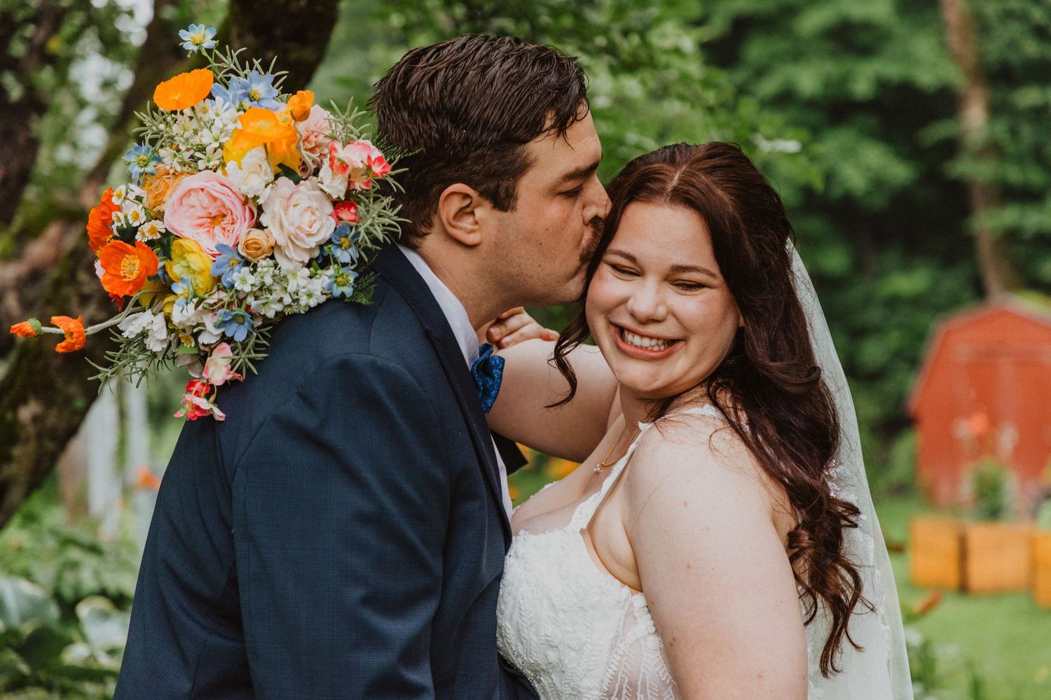 Groom in navy suit kisses bride wearing white gown and veil, holding colorful bouquet outdoors.