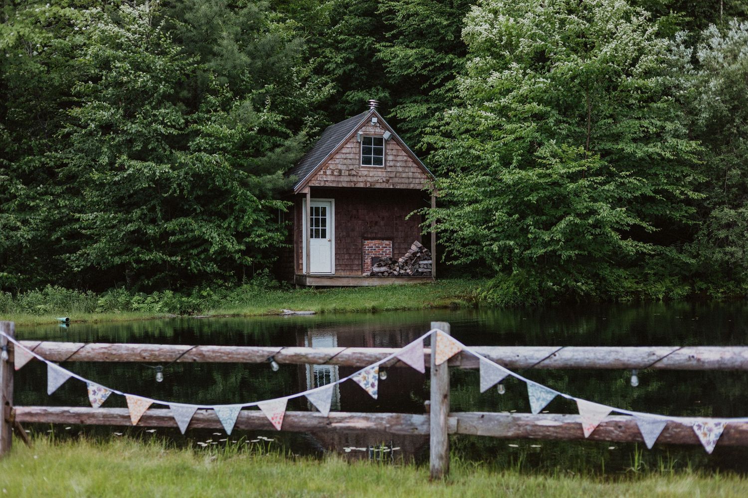 Rustic wooden cabin by a pond surrounded by lush trees, with colorful bunting on a fence in a serene countryside setting.