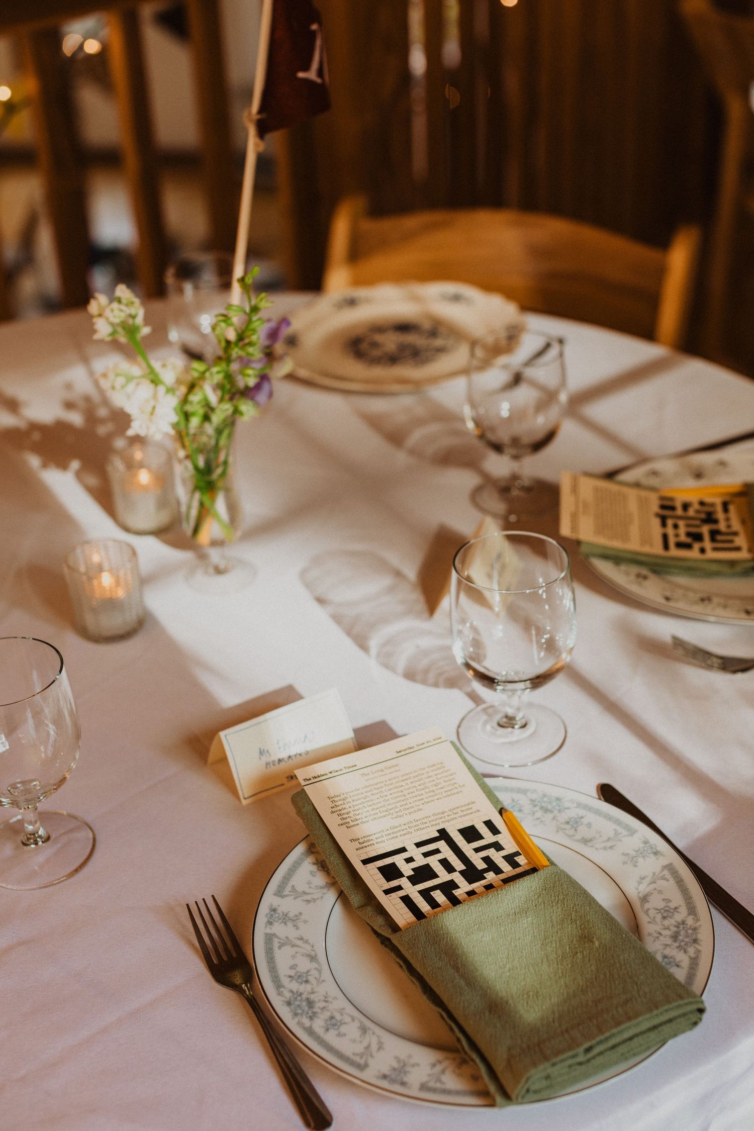 Elegant wedding table setting with crossword puzzle menu, green napkin, vintage china, wine glasses, and wildflower centerpiece.