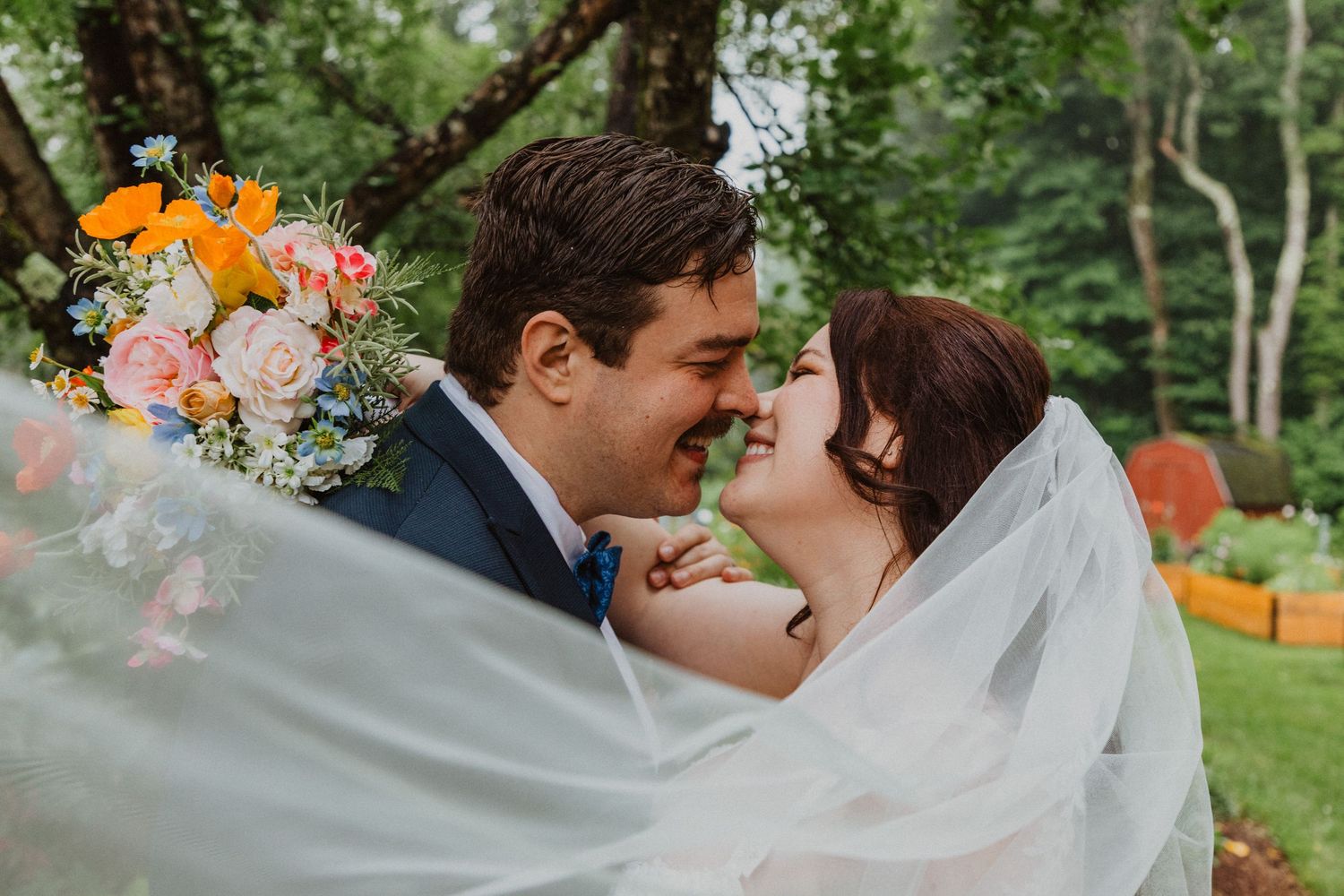Bride and groom sharing a romantic moment under flowing veil with colorful bridal bouquet.